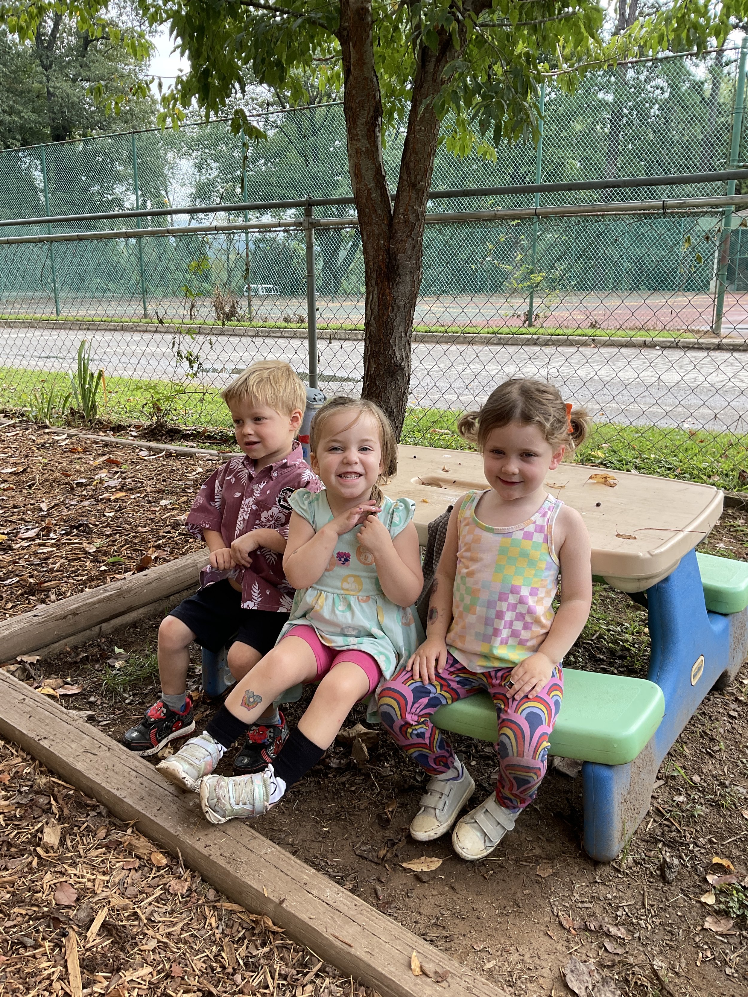 Three young children sitting on a colorful park bench with a small table, outdoors near a fenced tennis court and a tree, on a cloudy day.