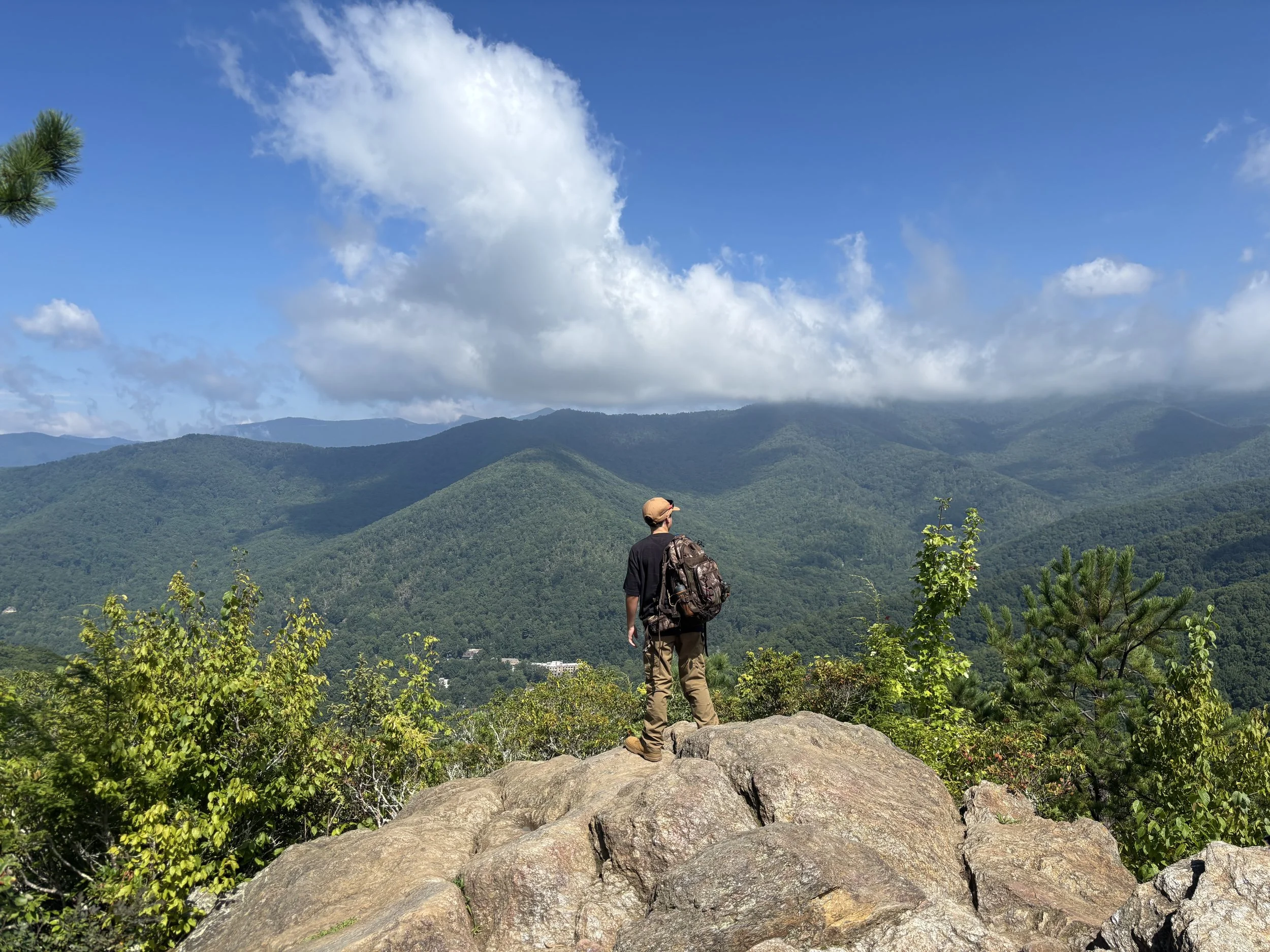 A man with a backpack standing on a large rock, looking at green mountains under a partly cloudy sky.