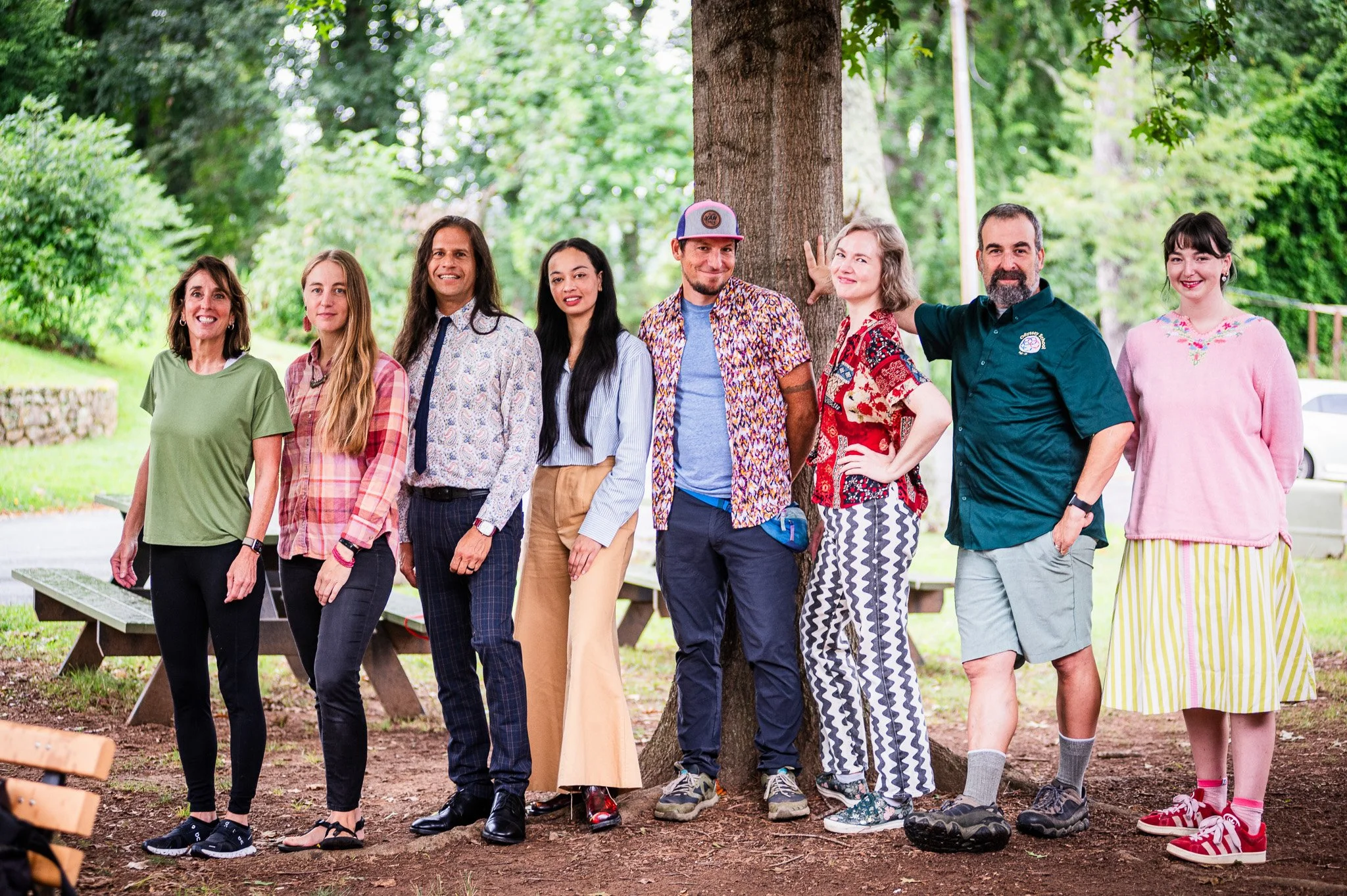 Group of nine diverse people standing outdoors in front of a large tree, smiling and posing for the photo in a park-like setting.
