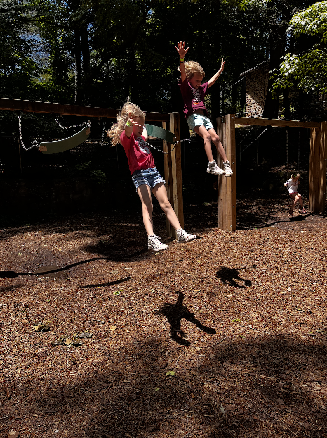 Two young girls are playing on a wooden playground in a wooded area. They are jumping off a structure, creating their shadows on the ground. Another girl is seen in the background on the playground.