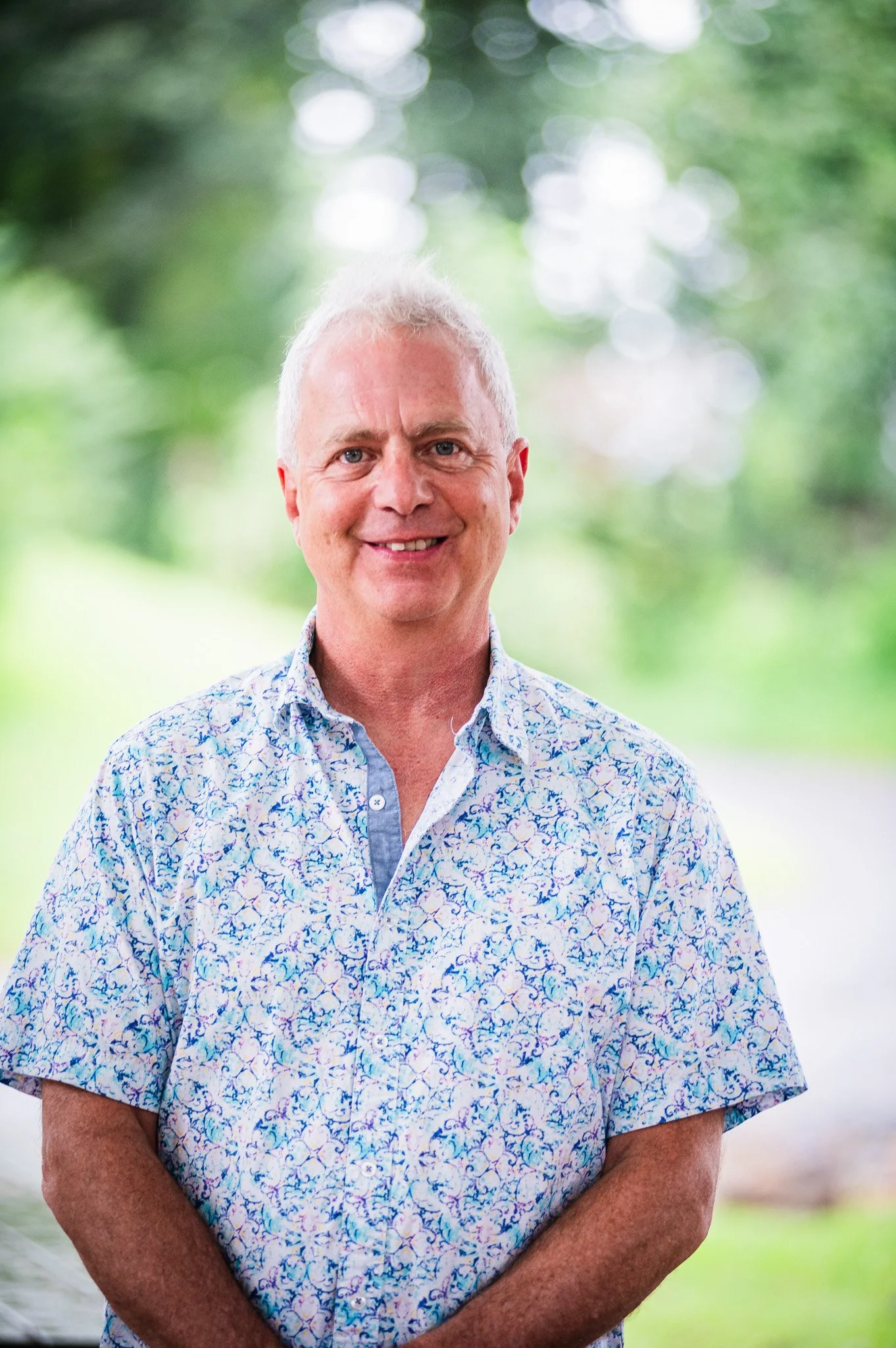 A smiling man with short gray hair wearing a short-sleeved, button-up shirt with a blue and white floral pattern, standing outdoors in front of a blurred background of green trees and sunlight.