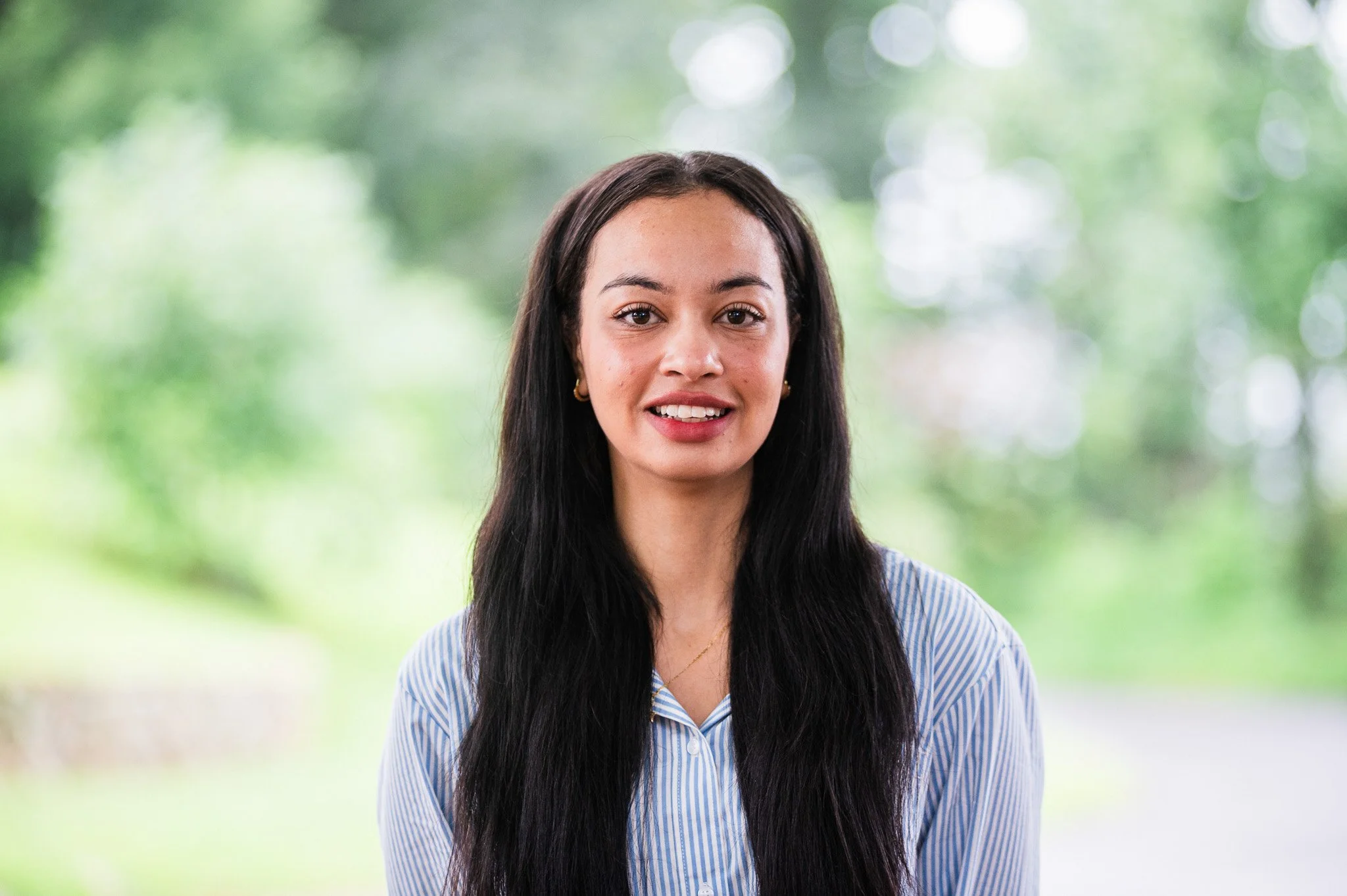 A woman with long dark hair, wearing a blue and white striped shirt, standing outdoors with a blurred green background.