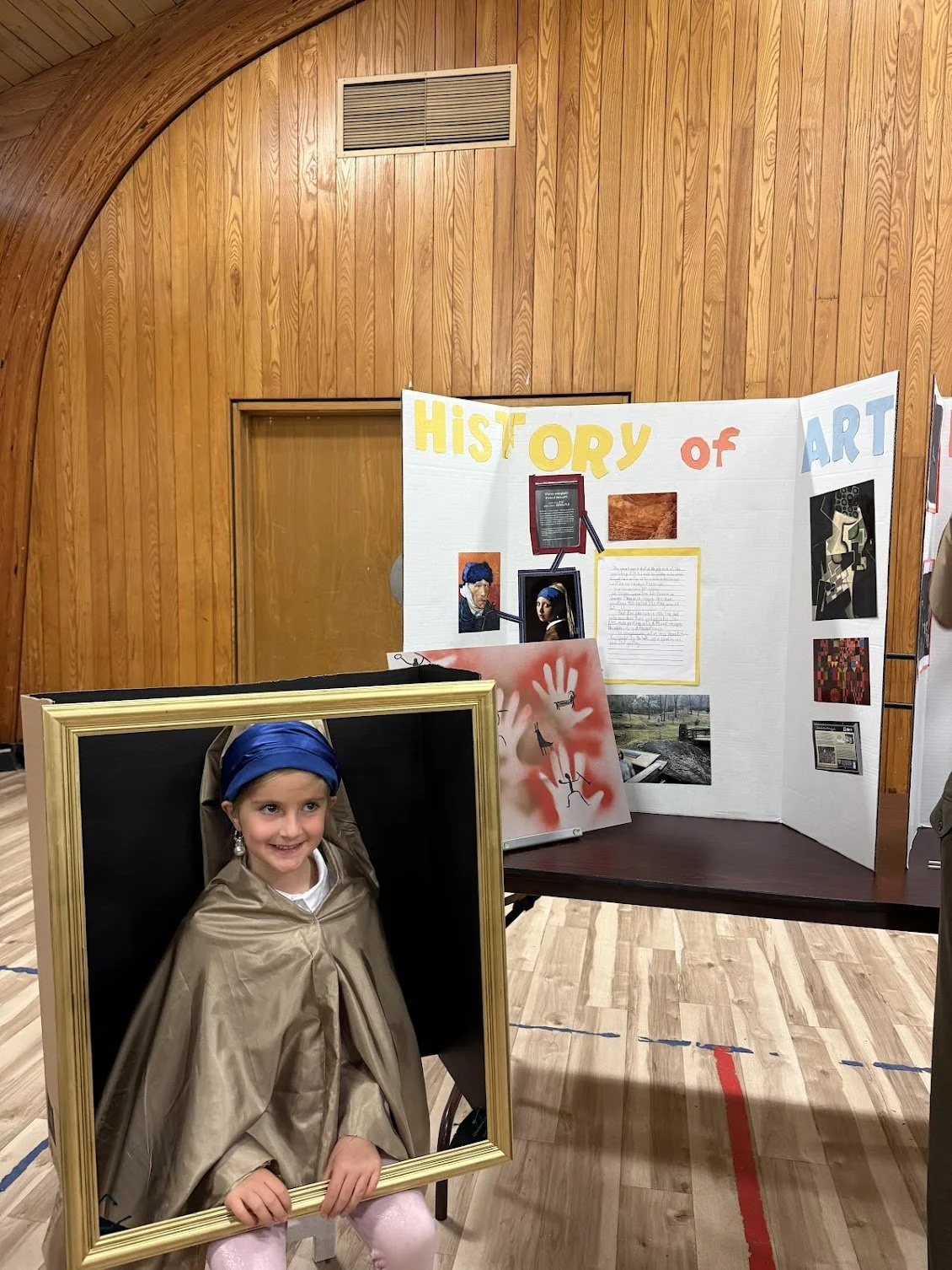 A young girl smiling and sitting in a large empty picture frame. She is wearing a blue headscarf and a beige cape. Behind her is a display board titled 'History of Art' with various images and text, set against a wood-paneled wall in a gymnasium or auditorium.