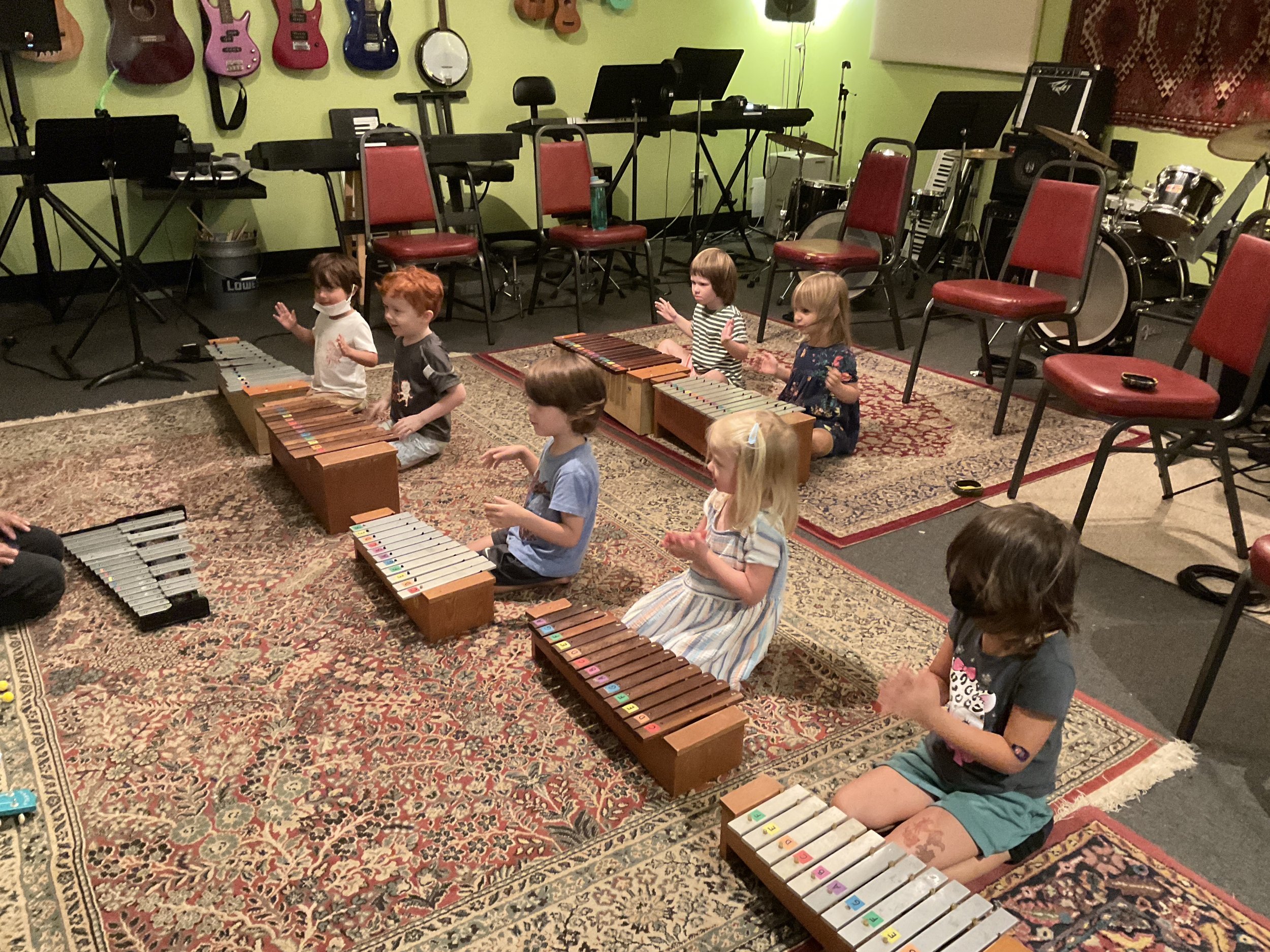 Children sitting cross-legged on a carpet playing xylophones in a music classroom with musical instruments on the wall and chairs in the background.