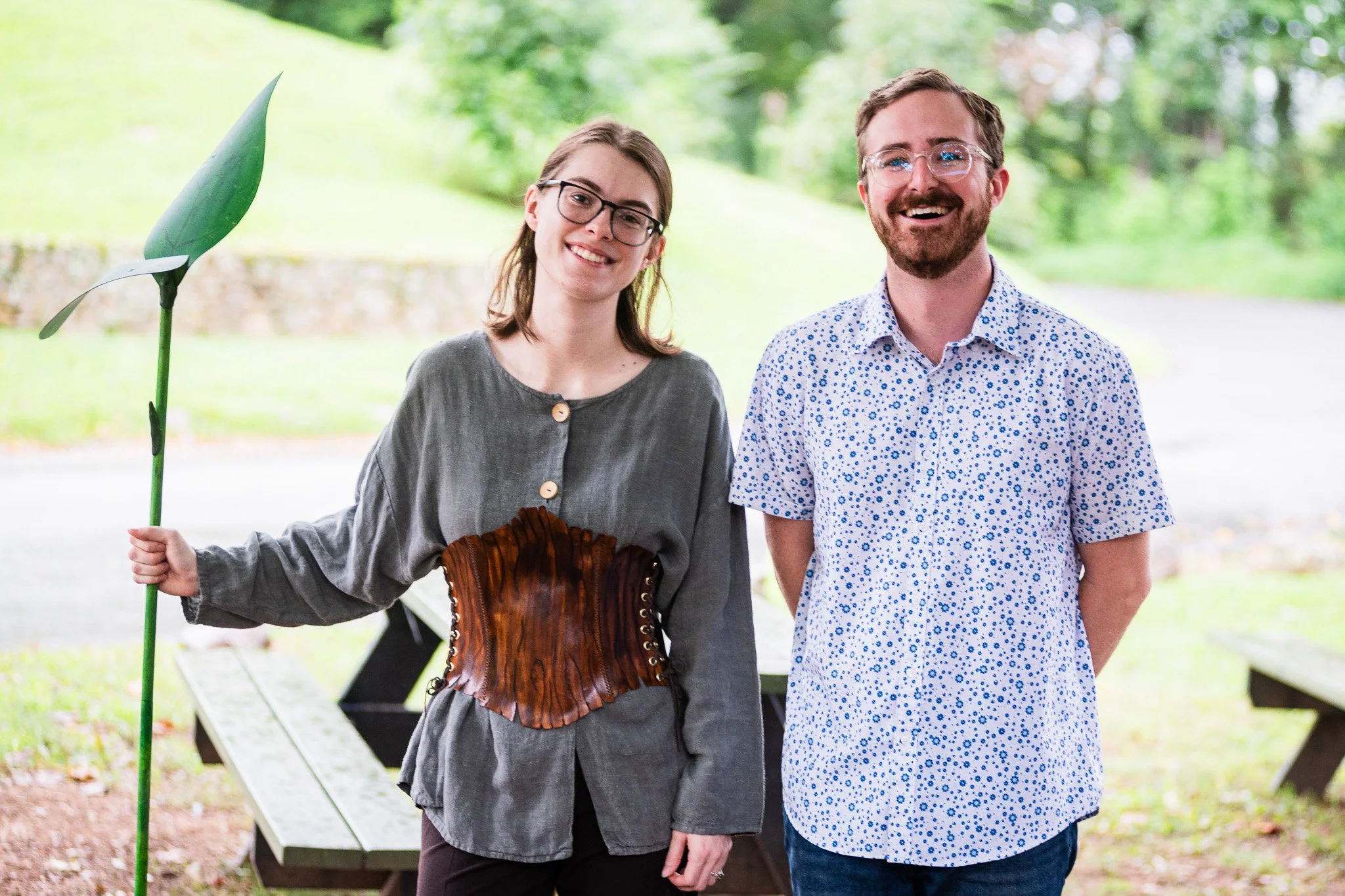 A smiling woman and man standing outdoors in a park, with the woman holding a decorative green stem with leaves. The woman is wearing glasses, a gray blouse, and a brown leather corset, while the man is wearing glasses, a white shirt with blue floral pattern, and has a beard. They are standing behind a wooden picnic table with a grassy background and trees.
