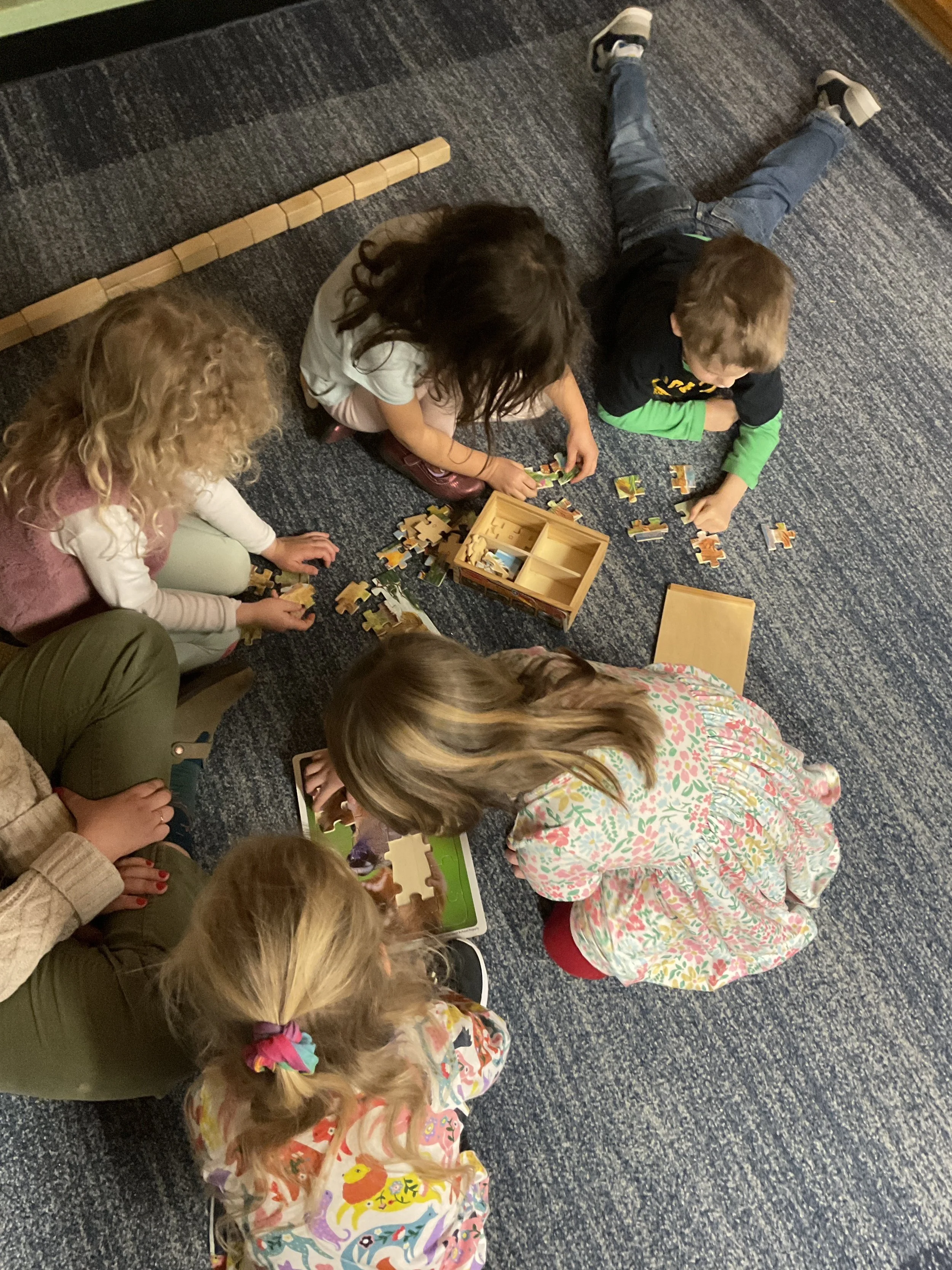 Five preschool children sitting on a blue carpeted floor playing with puzzles, some using a tablet, with a wooden puzzle board also visible.