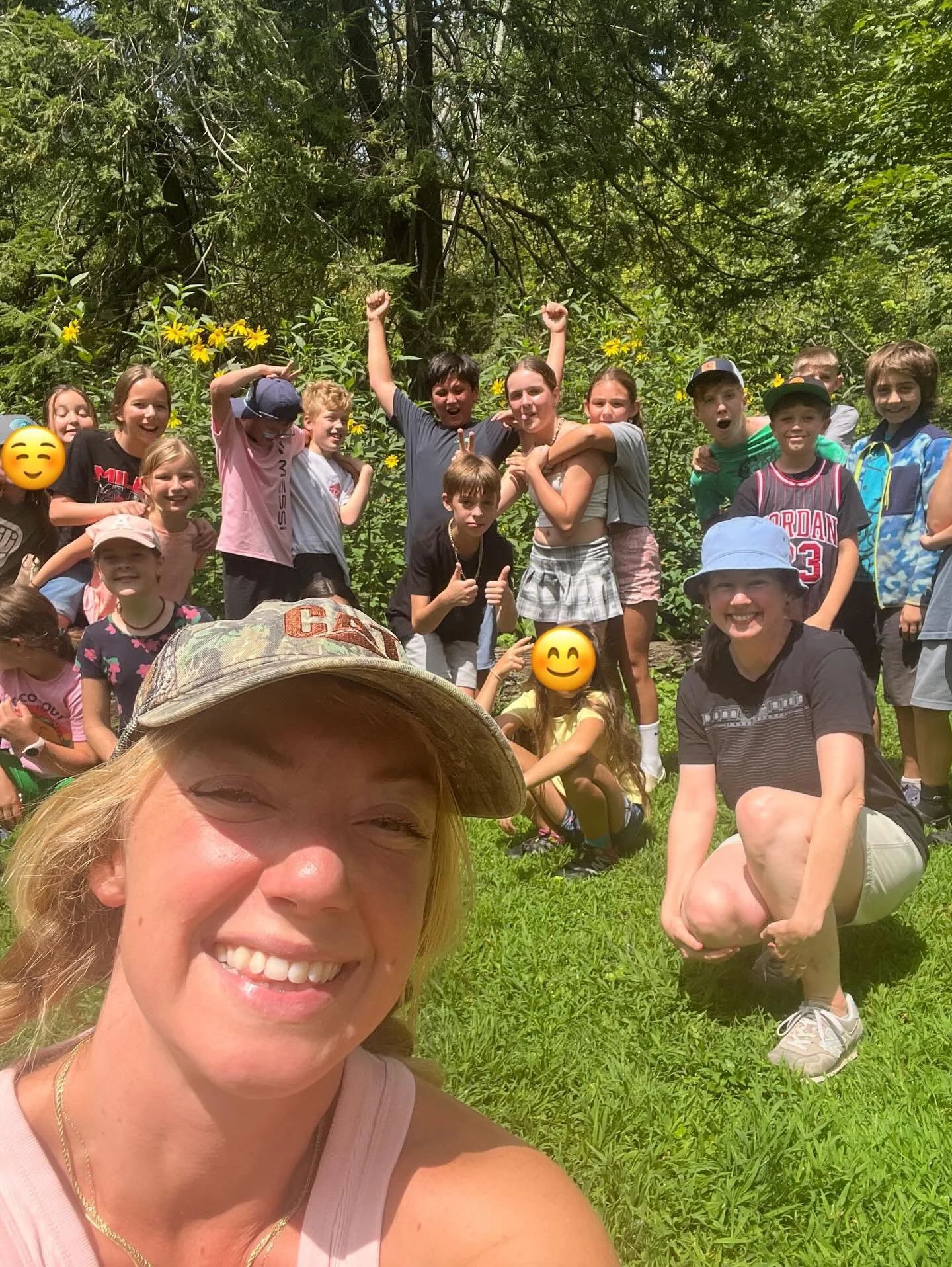 A group of children and two women outdoors on a sunny day, posing for a group photo near lush green trees and yellow flowers.