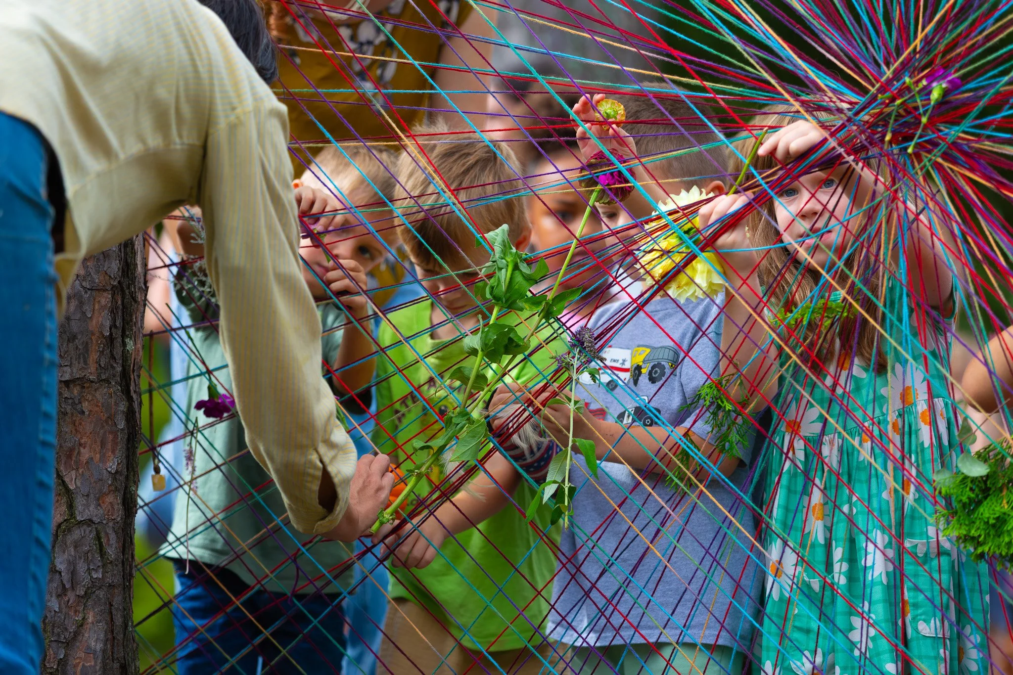 Group of children behind a colorful string web, looking through and reaching for plants and flowers, outdoors.