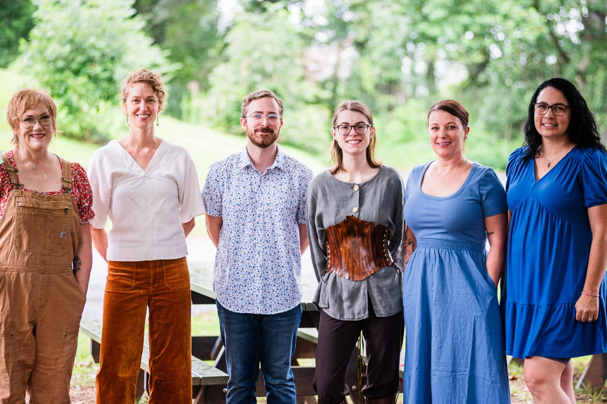 Group of six women and one man standing outdoors in a park, smiling at the camera.