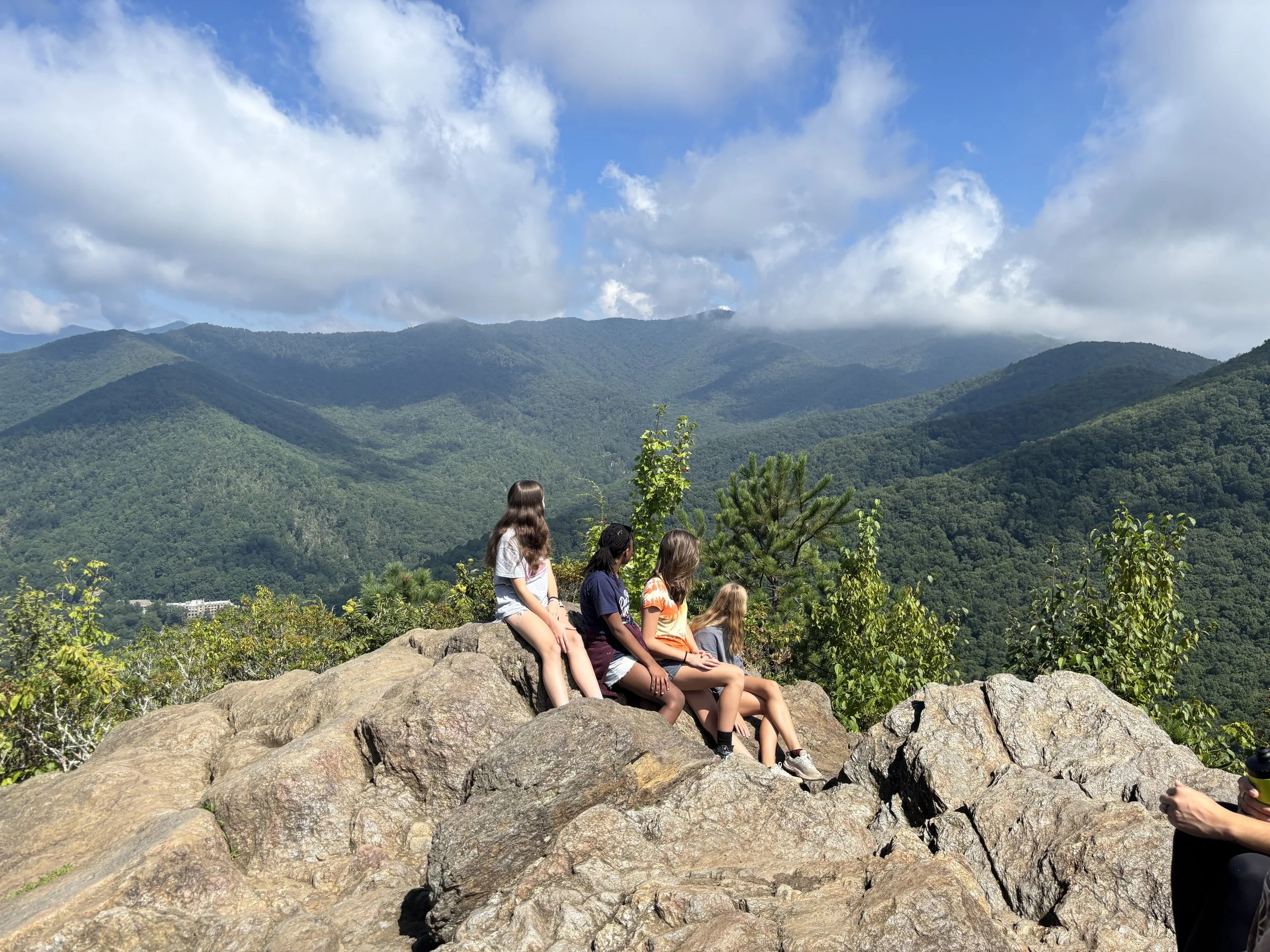 Four girls sitting on rocks overlooking a the blue ridge mountain range with clouds in the sky.