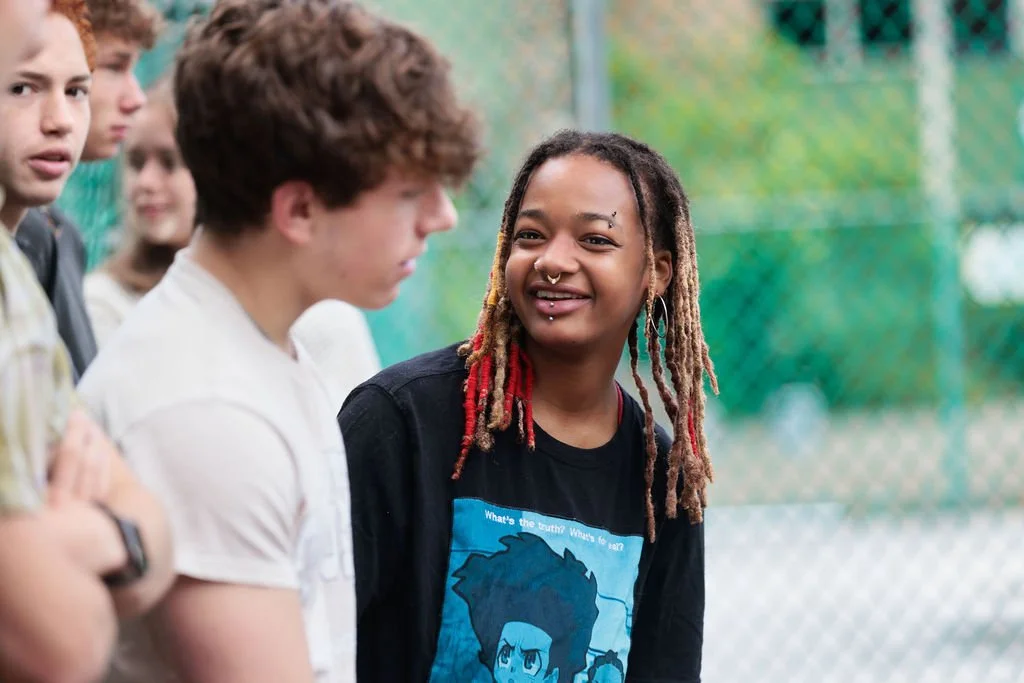 A young woman with dreadlocks and piercings smiling and talking to a group of teenagers outside near a chain-link fence.