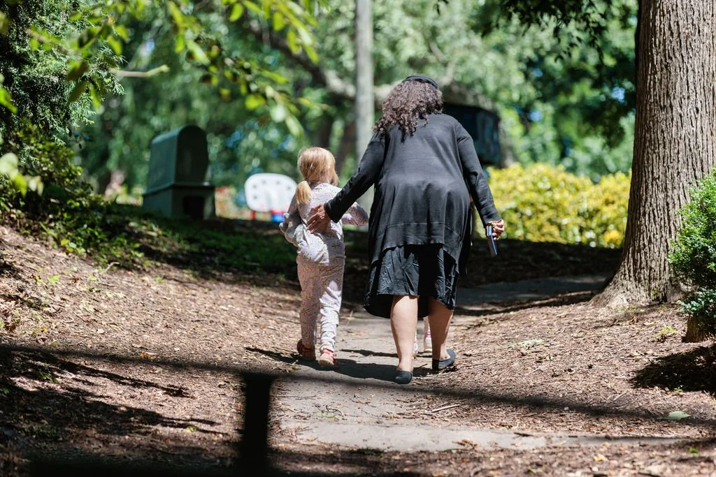 A woman and a young girl walking hand in hand on a dirt path through a wooded park.