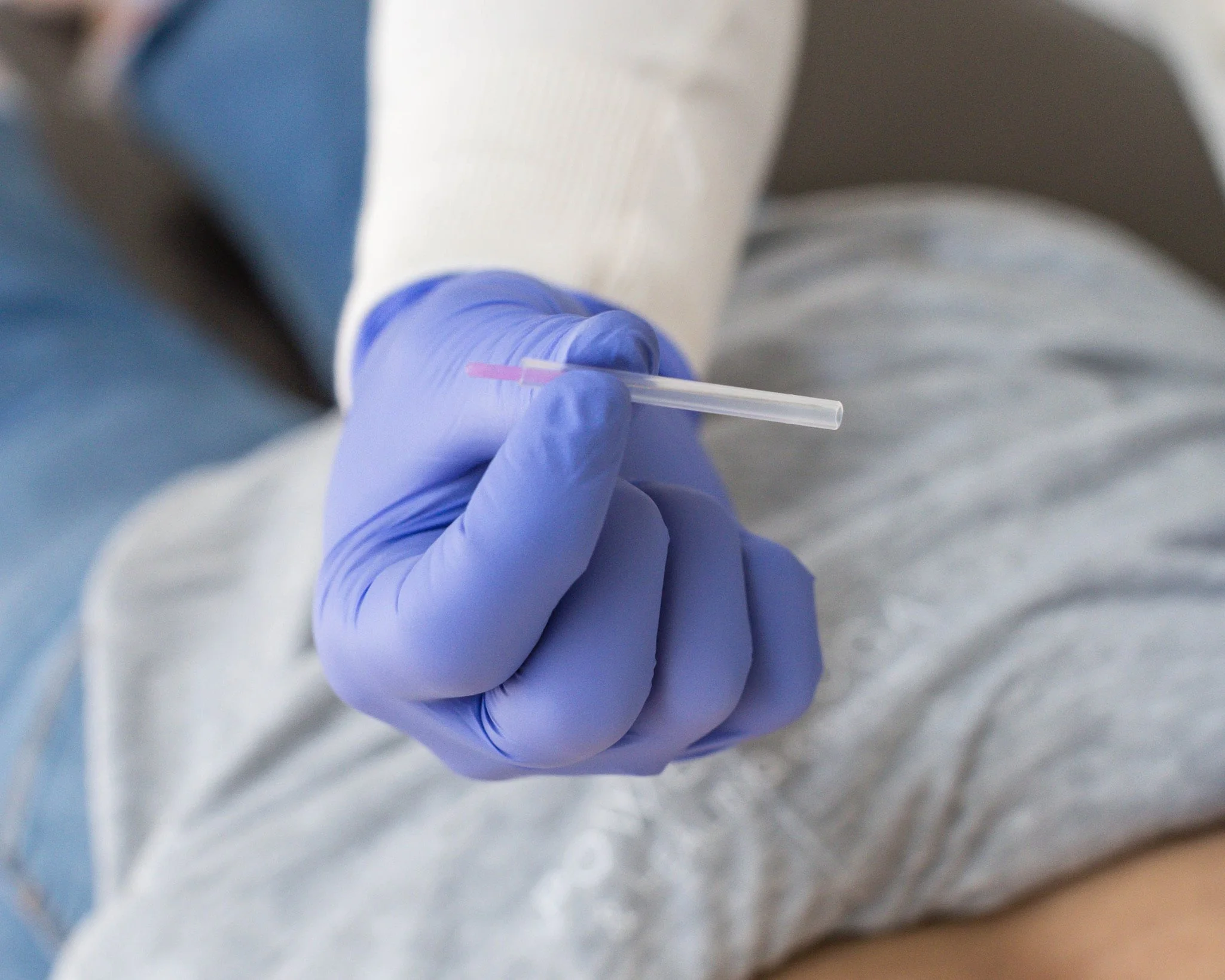 A person wearing purple gloves holding a small needle, with part of their arm visible resting on a gray surface.