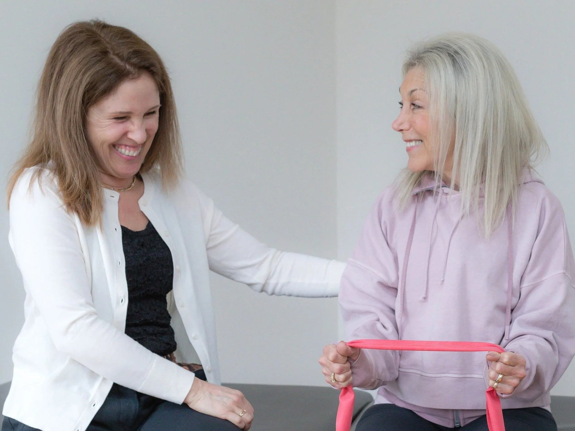 Two women smiling and interacting, one holding a pink resistance band, in a plain indoor setting.