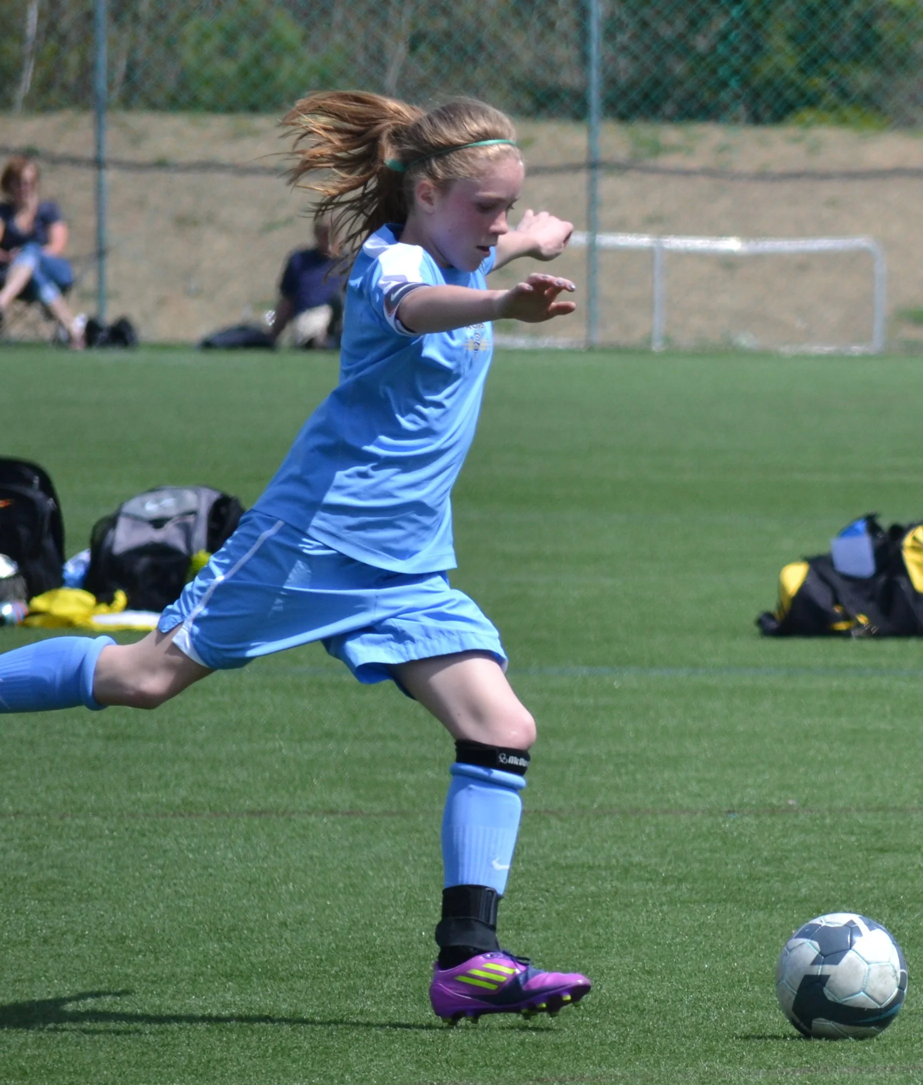 A young girl in a blue soccer uniform is running on a soccer field, about to kick a soccer ball. She has long hair tied back and is wearing purple cleats and knee-high socks.