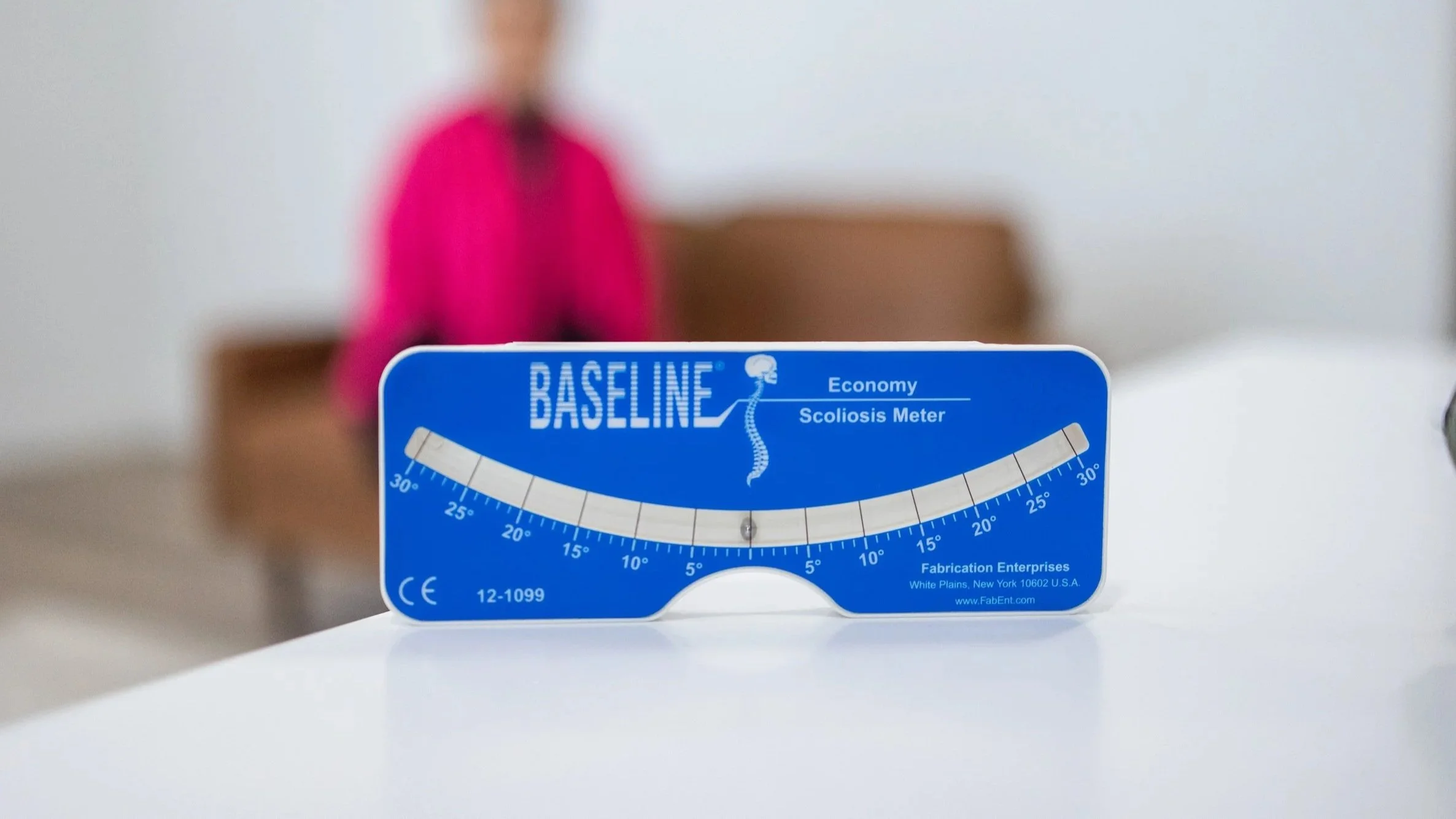 A blue scoliosis meter titled 'Baseline' with a scale from -30 to +30 degrees, and a figure with a spine illustration, placed on a white surface. A blurred person stands in the background.