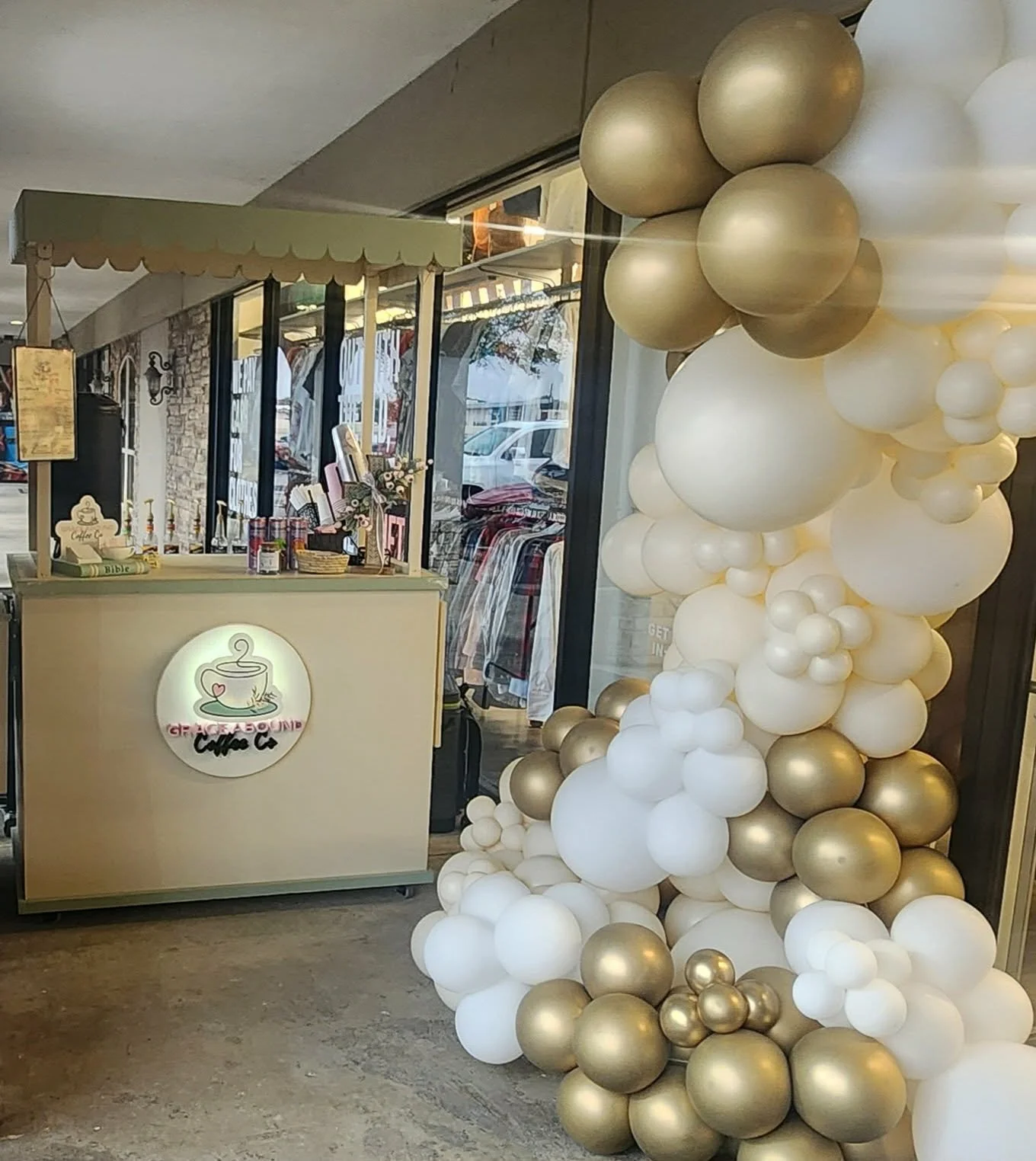 Decorative balloon arch featuring white, gold, and champagne-colored balloons next to a coffee shop counter with a sign that says 'Coffee Co'. The cafe has a light wood counter with a cupcake decoration and various items on top, located inside a shopping mall or retail space with clothing racks visible in the background.