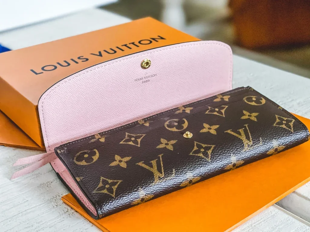 Portafoglio Louis Vuitton con motivo monogramma marrone e dettagli rosa, aperto con scatola e scatole arancioni sullo sfondo.