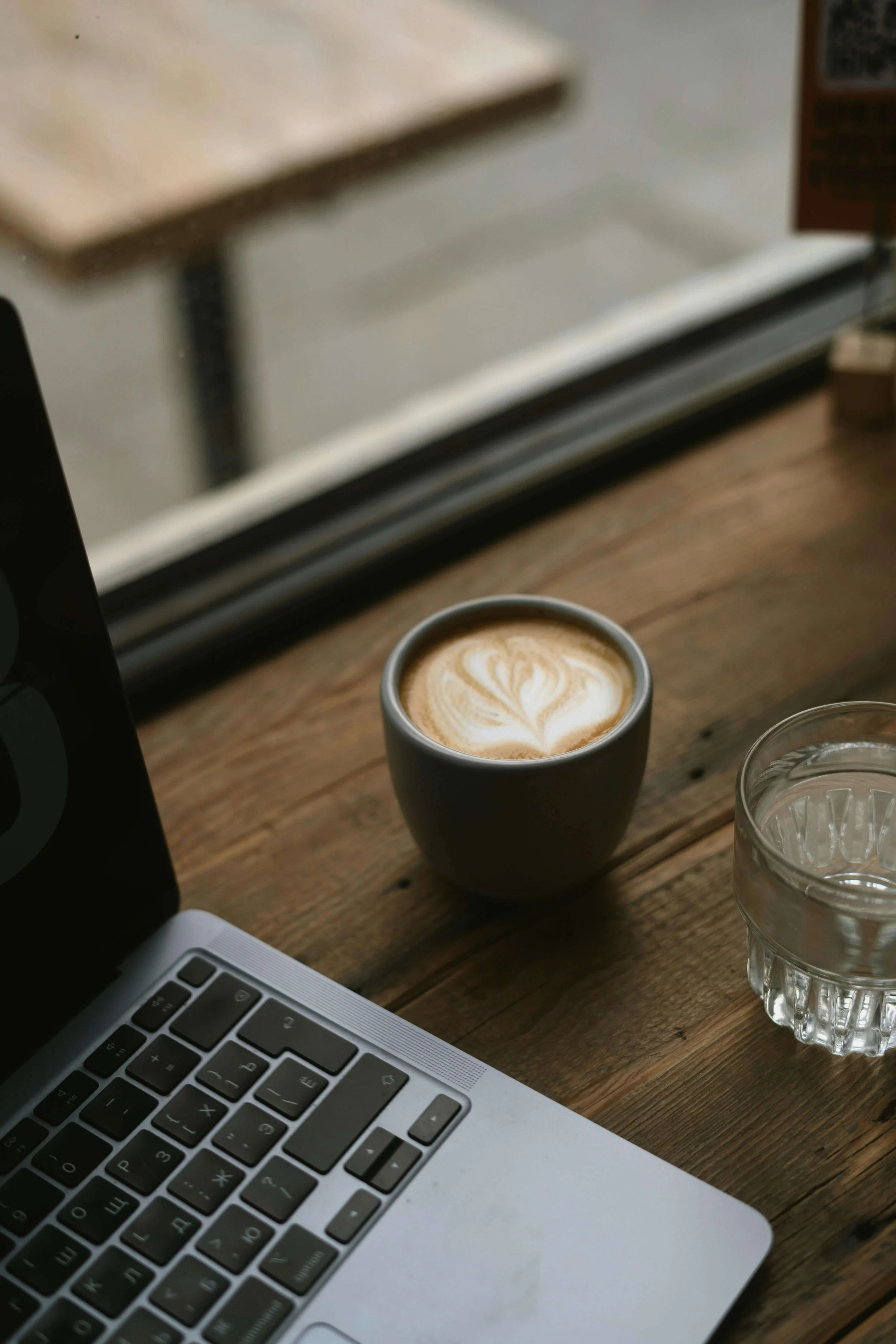 A wooden table with a silver laptop showing a keyboard, a glass of water, a cappuccino with latte art, a black object, and part of a menu or book.