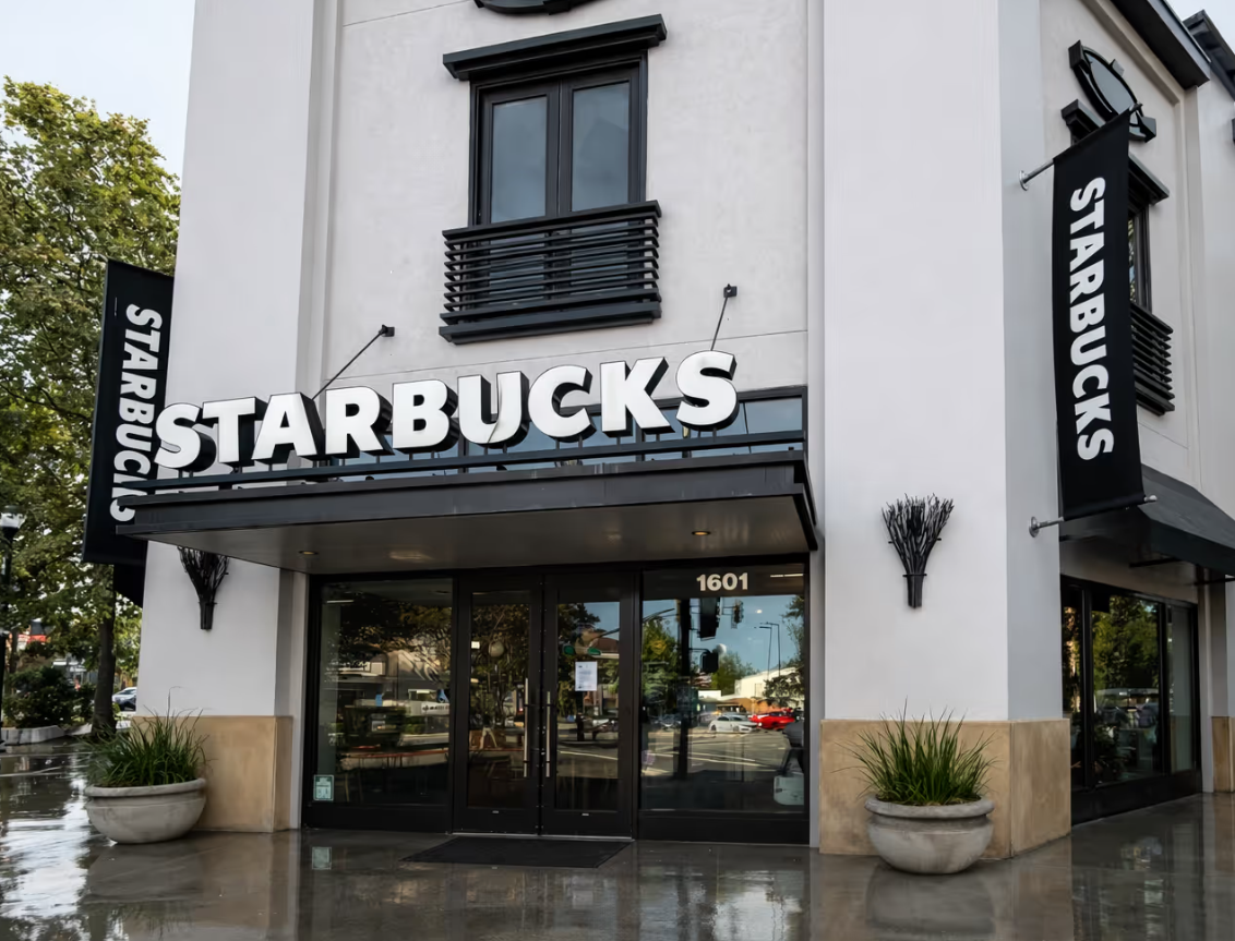 Exterior view of a Starbucks store with large white signage, black banners on sides, and two potted plants outside.