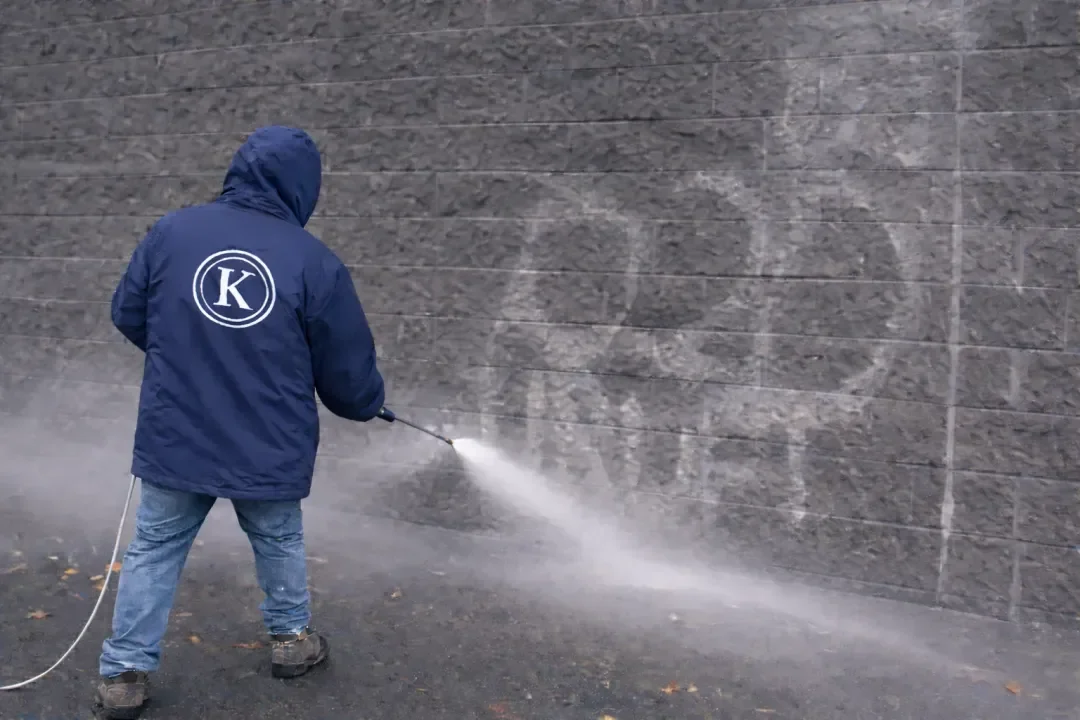 Person wearing a blue jacket with a circular logo spray cleaning a large wall with a pressure washer, standing on a gravel surface.