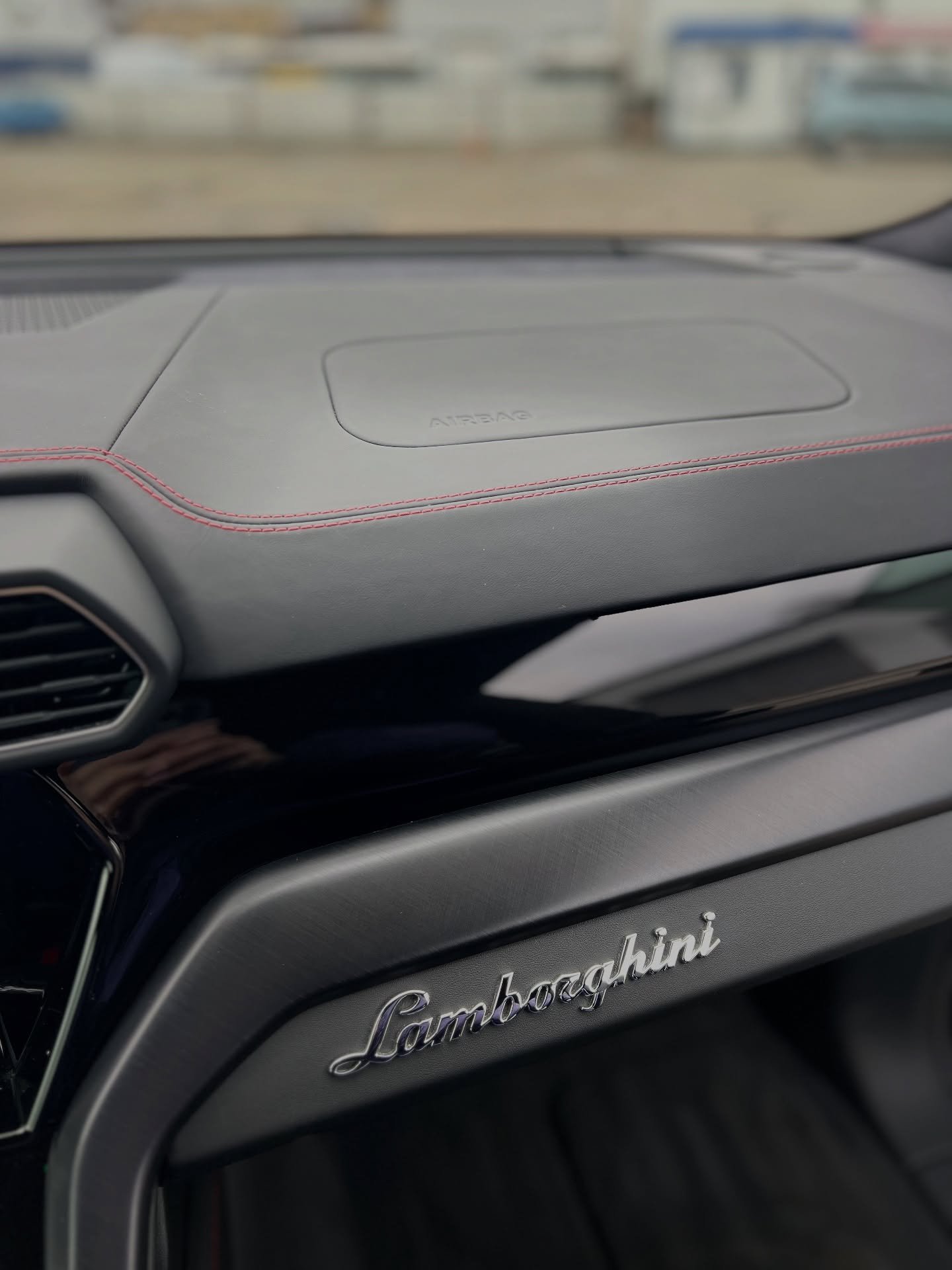 Close-up of the dashboard interior of a Lamborghini, showing the Lamborghini logo and part of the air vent, with a blurred view of the outside through the windshield.