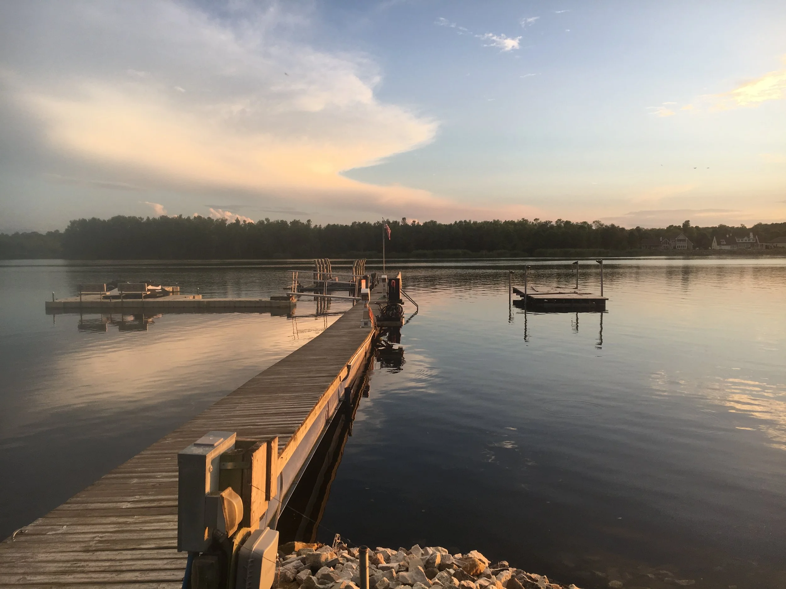 A wooden dock extends into a calm body of water, with a flagpole displaying an American flag. The sky above is partly cloudy with a soft pastel sunset or sunrise glow, and trees and houses are visible along the shoreline in the background.