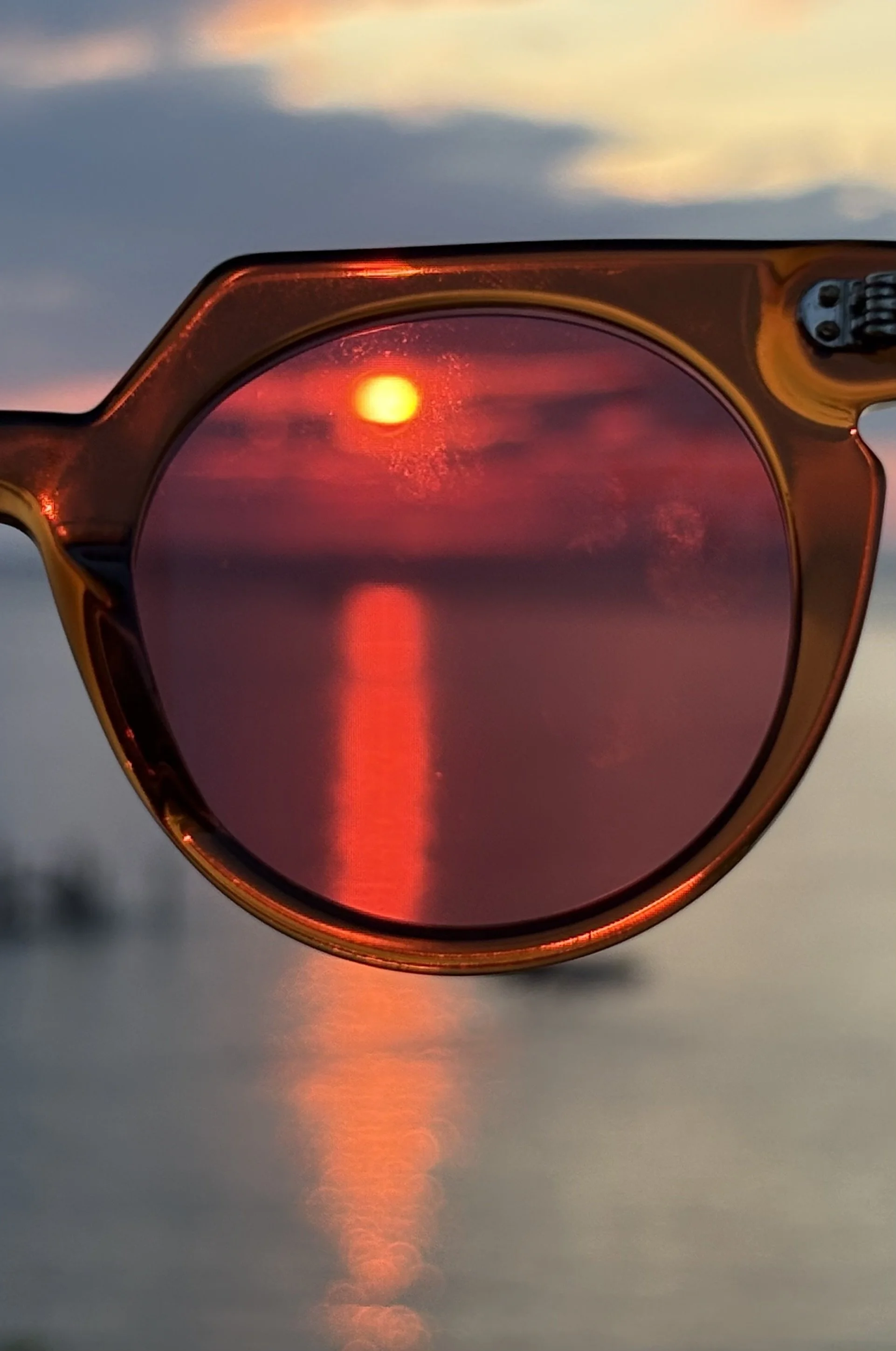 A pair of sunglasses with reflective pinkish-red tinted lenses, capturing the reflection of the setting sun over a body of water at sunset.