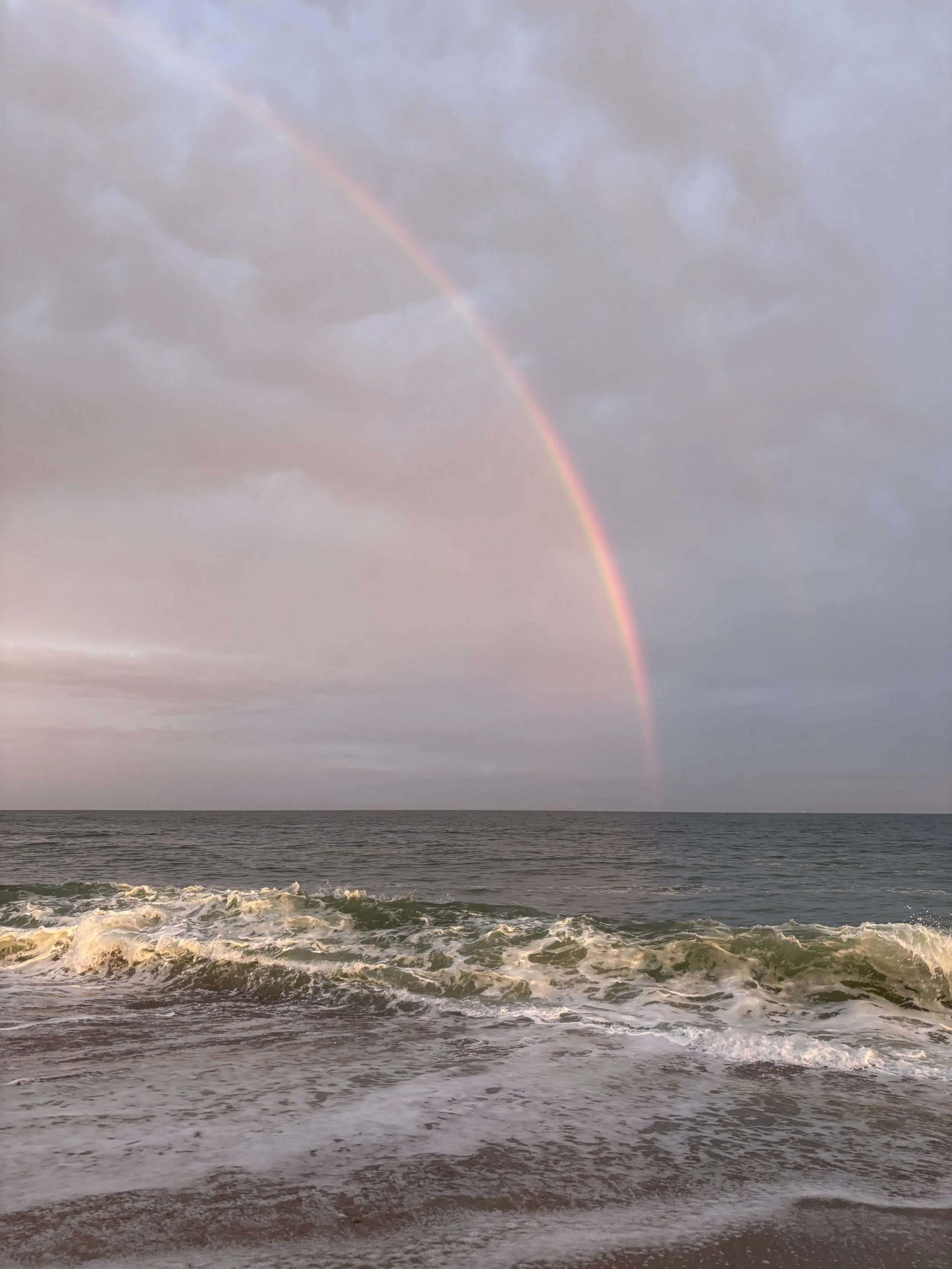 A rainbow arching over a choppy ocean with waves crashing onto the shore under a cloudy sky.