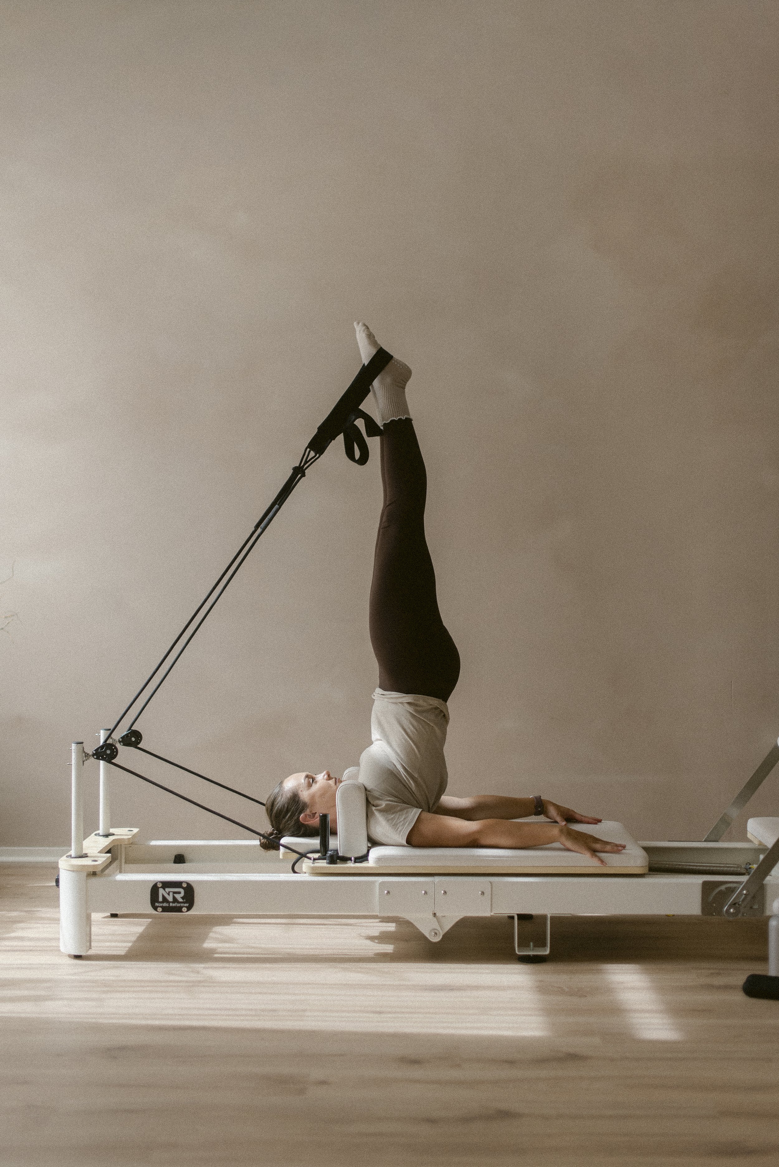 Woman performing Pilates on reformer machine, lying on back with legs extended vertically, in a room with wooden flooring and neutral walls.