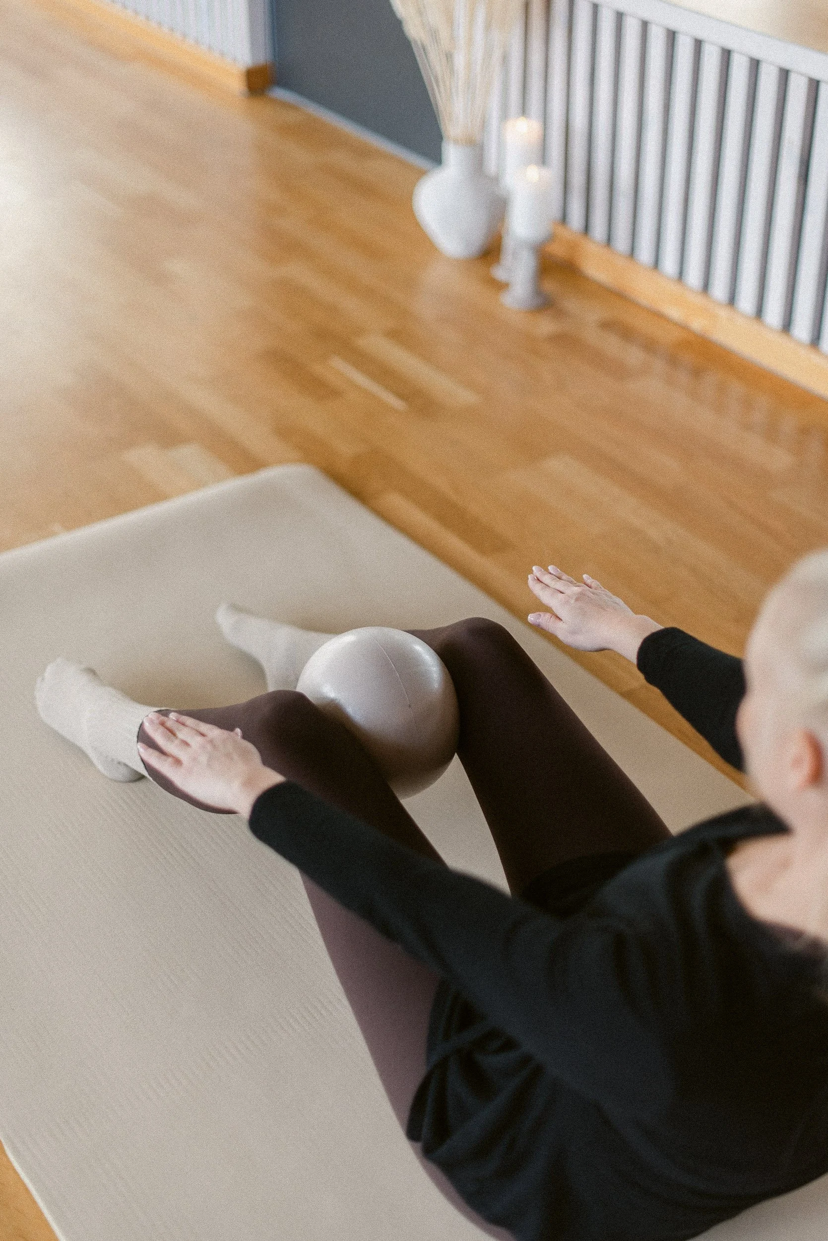 Person sitting on a beige exercise mat with their legs spread apart, balancing a gray exercise ball on their lower body, in a room with wooden floors and white decor.