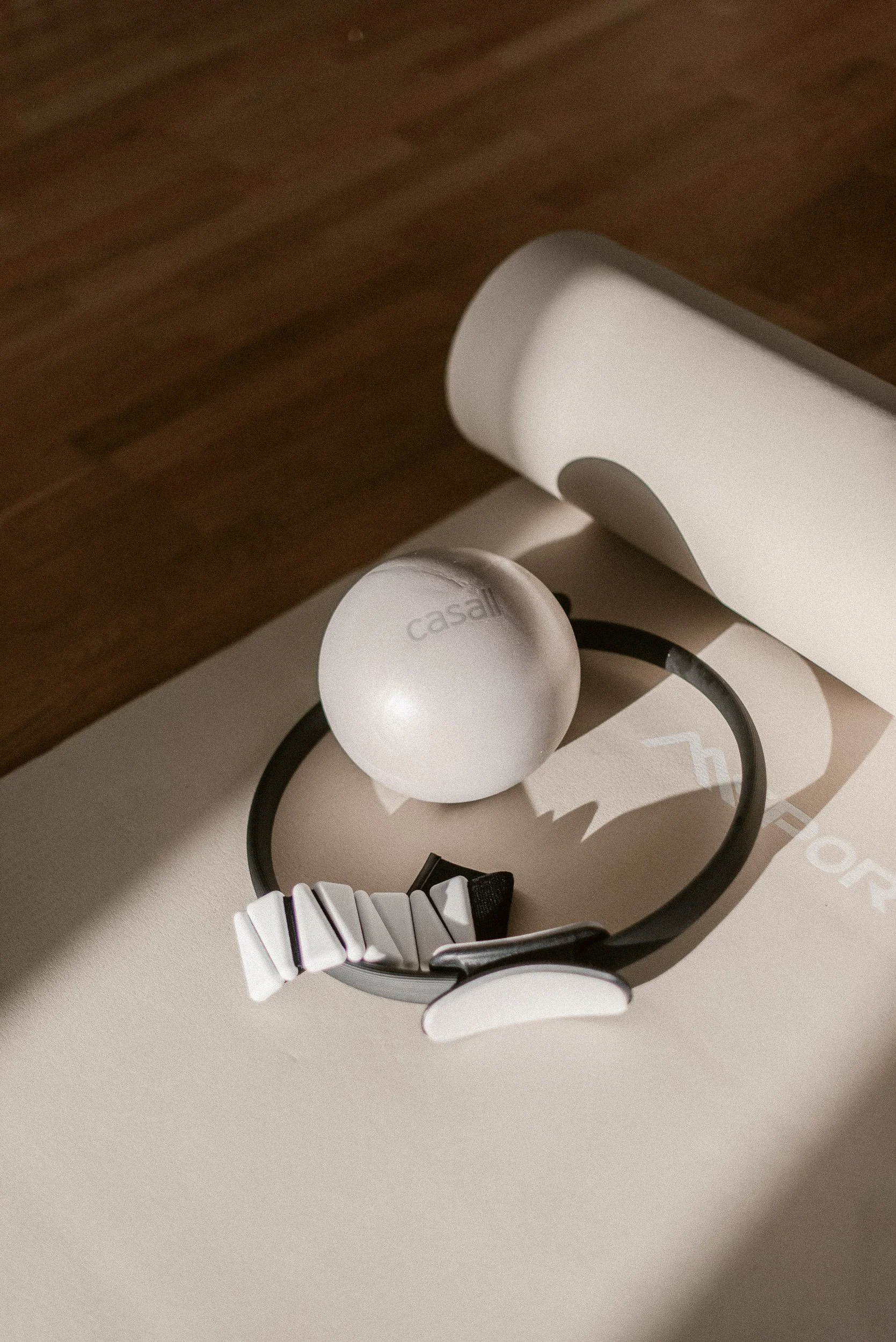 A ball with the word 'casali' on it, a black pilates ring, and ankle weights on a light-colored surface with a wooden floor beneath.