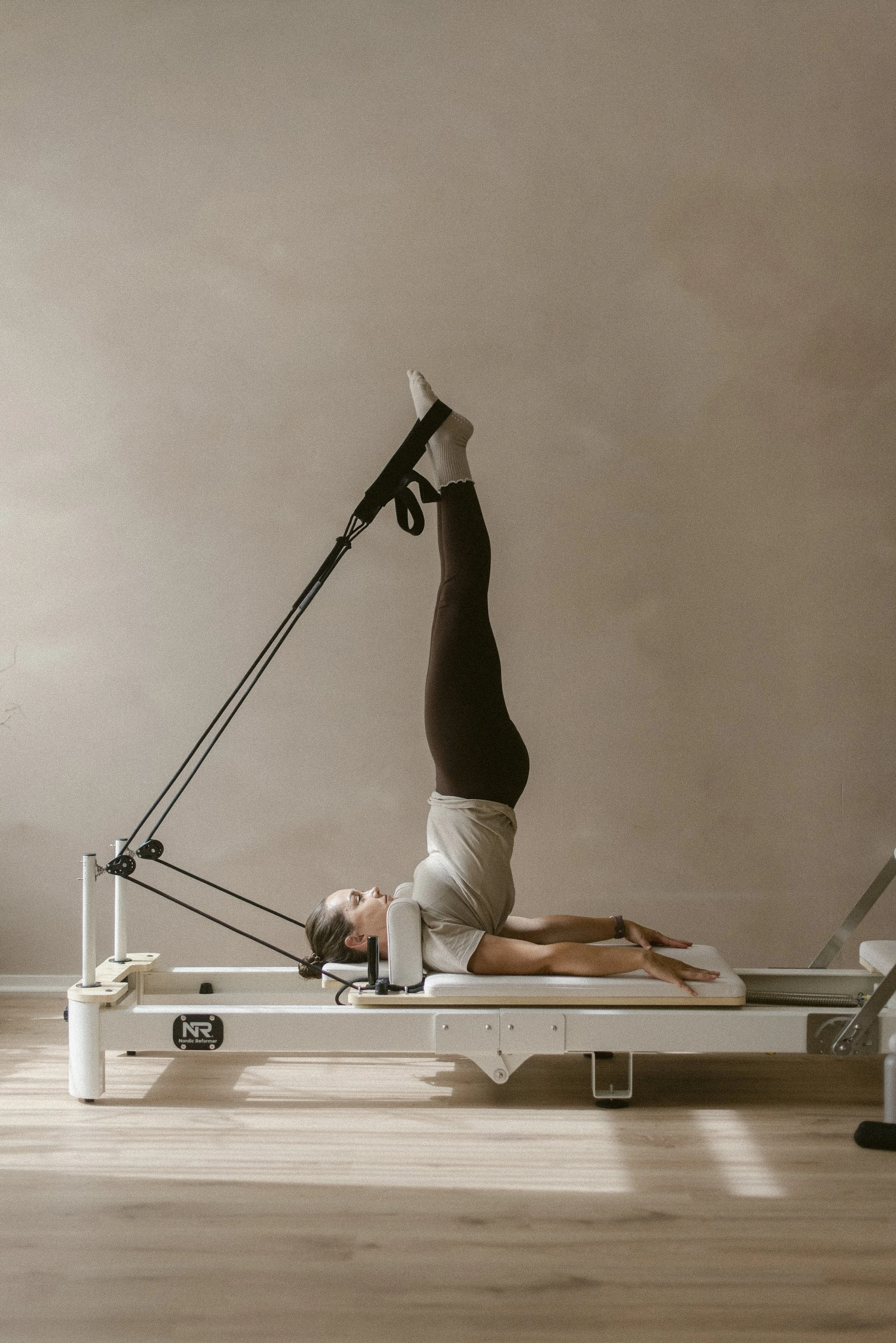 A woman practicing Pilates on a reformer machine in a room with plain beige walls and wooden flooring.