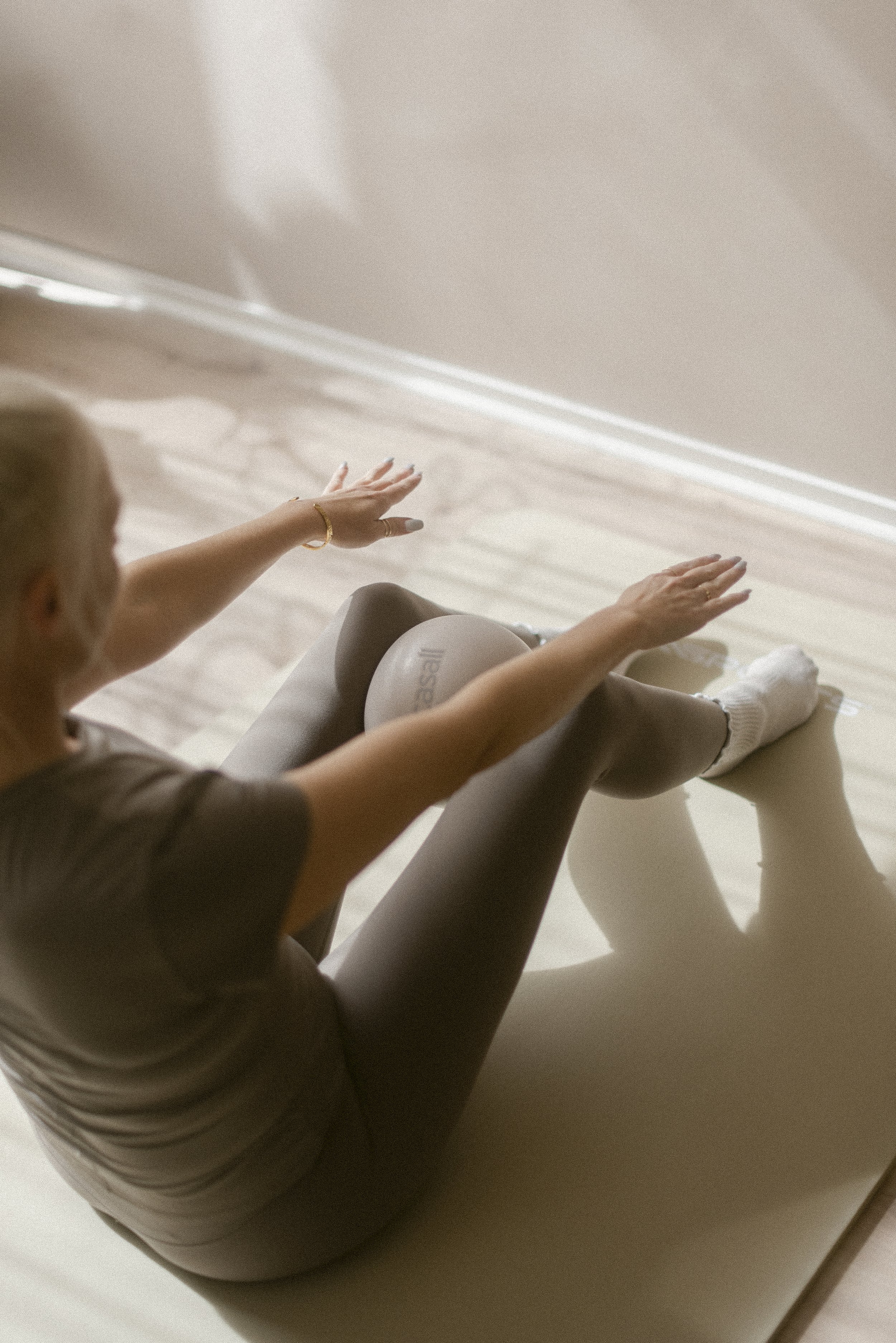 A person sitting on a pilates mat, wearing white socks and a short-sleeved shirt, reaching out with both arms towards a wall. The room has beige walls and light-colored wooden flooring.