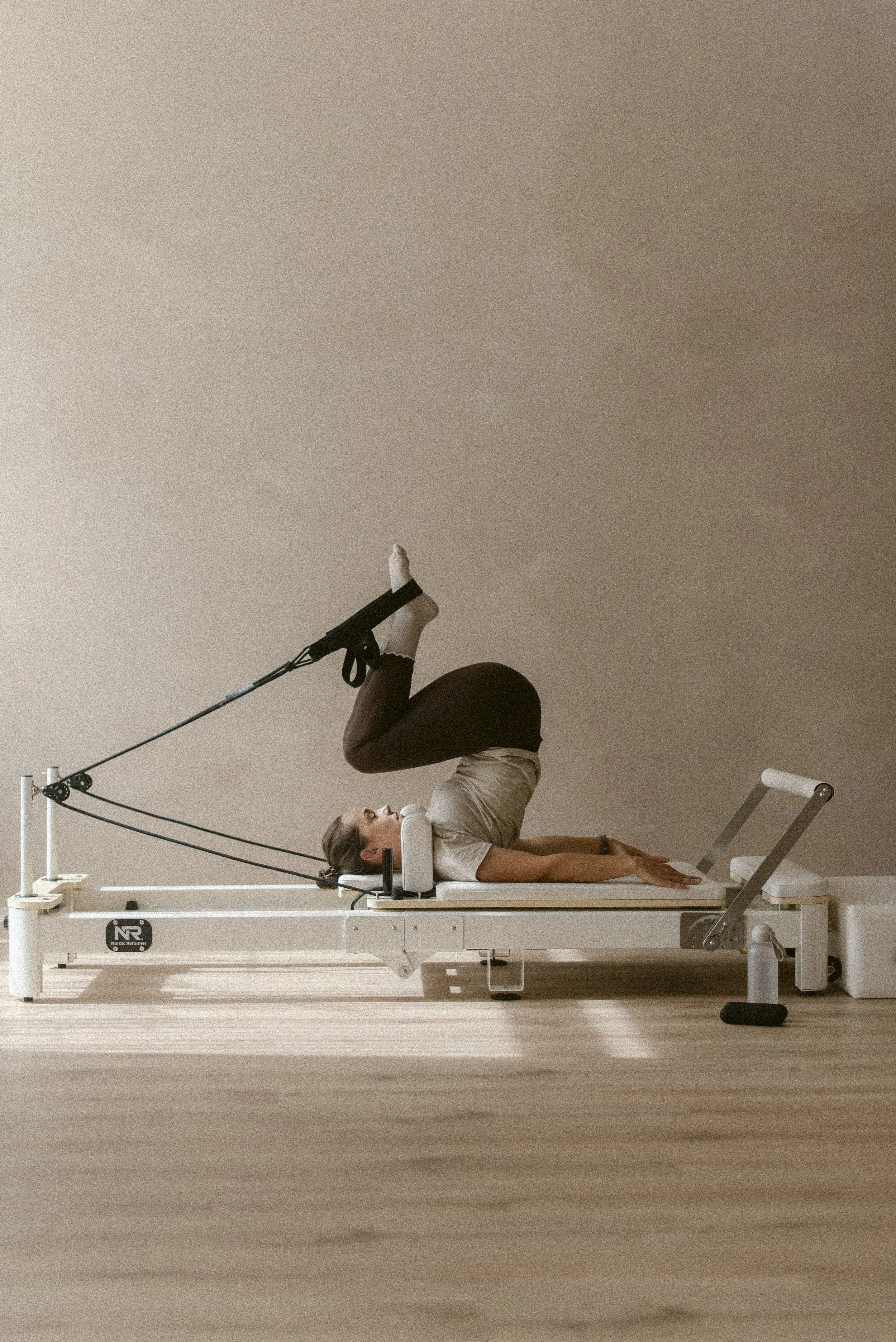 A woman lying on her back on a pilates reformer machine with her legs lifted and bent at the knees, and wearing compression stockings. The room has a beige wall and wood flooring.