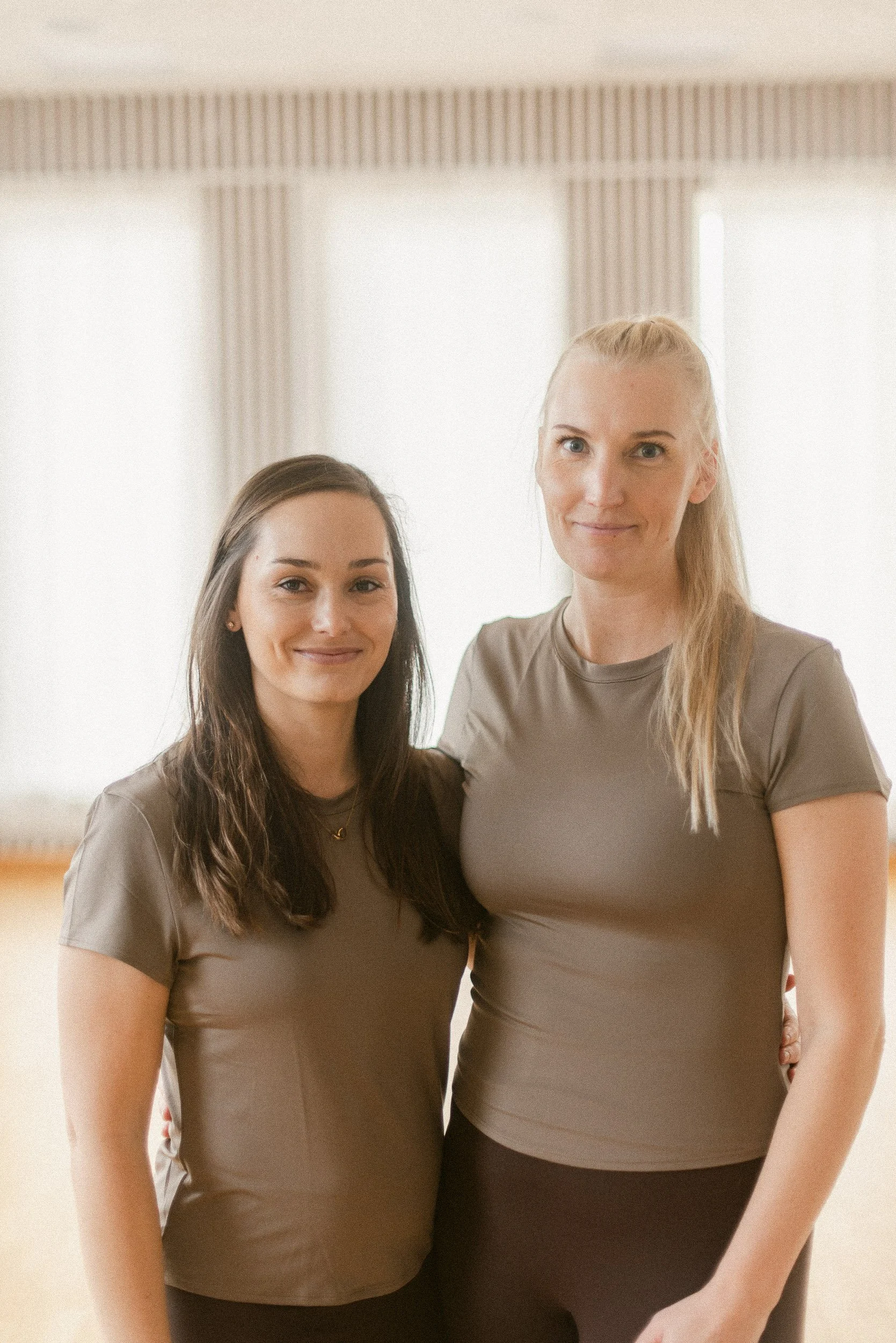 Two women standing close together in a room with bright windows and curtains, smiling at the camera.
