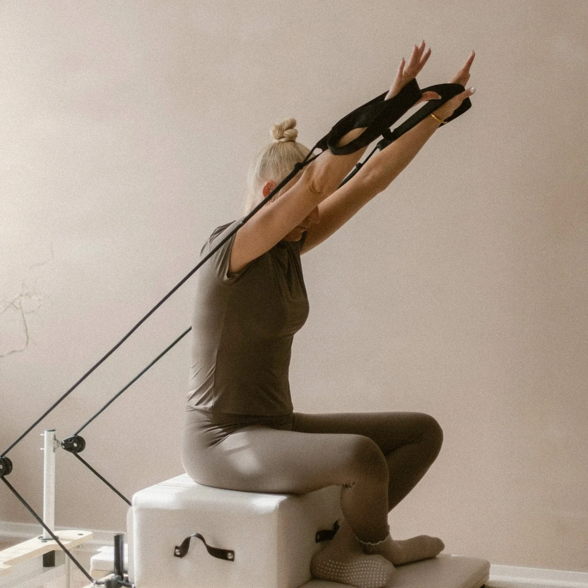 A woman with blonde hair in a bun practicing pilates in a kneeling pose on a white reformer against a plain beige wall.