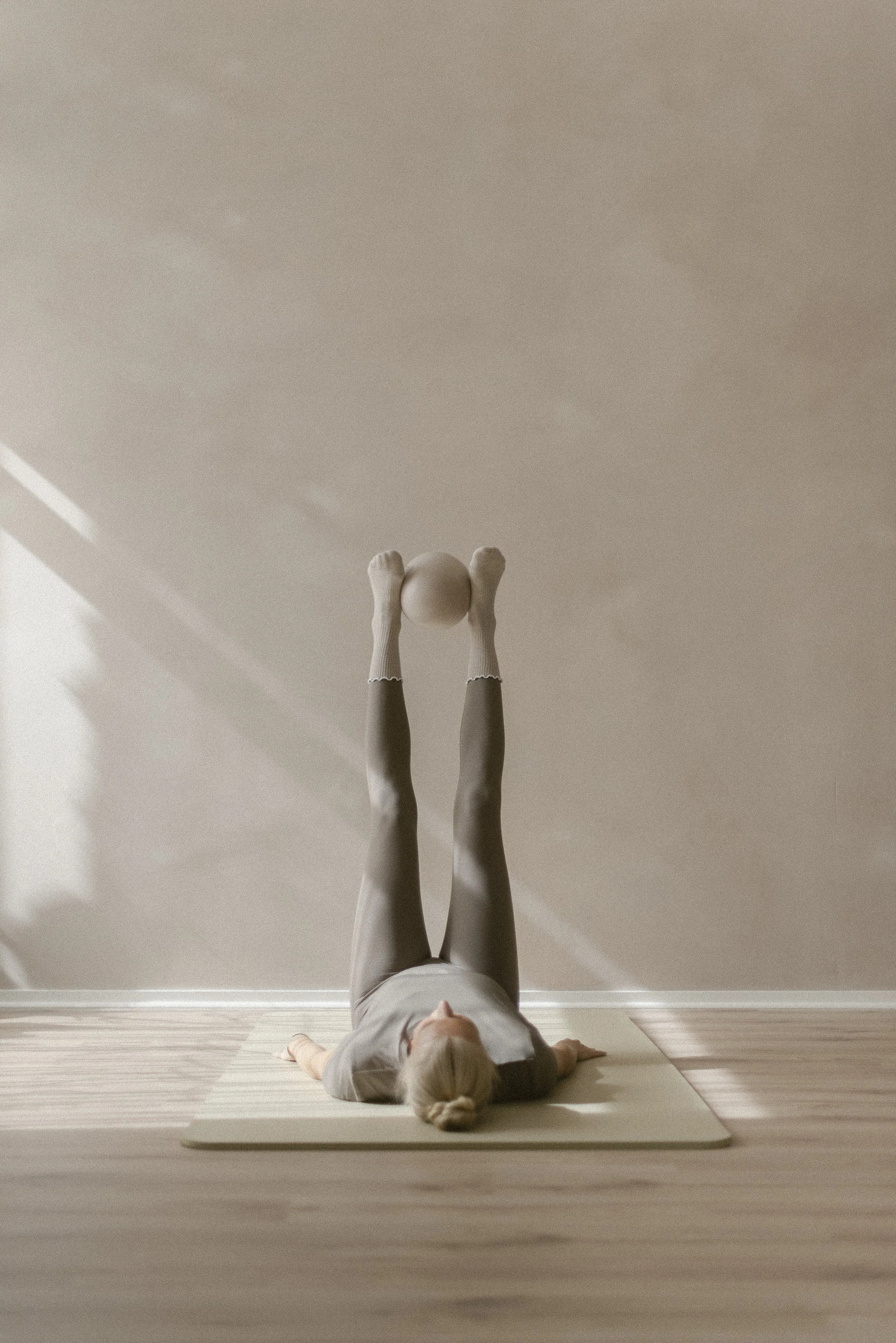 A person with blond hair, lying on their back on a pilates mat, performing a leg raise with legs extended upward and arms resting on the floor.