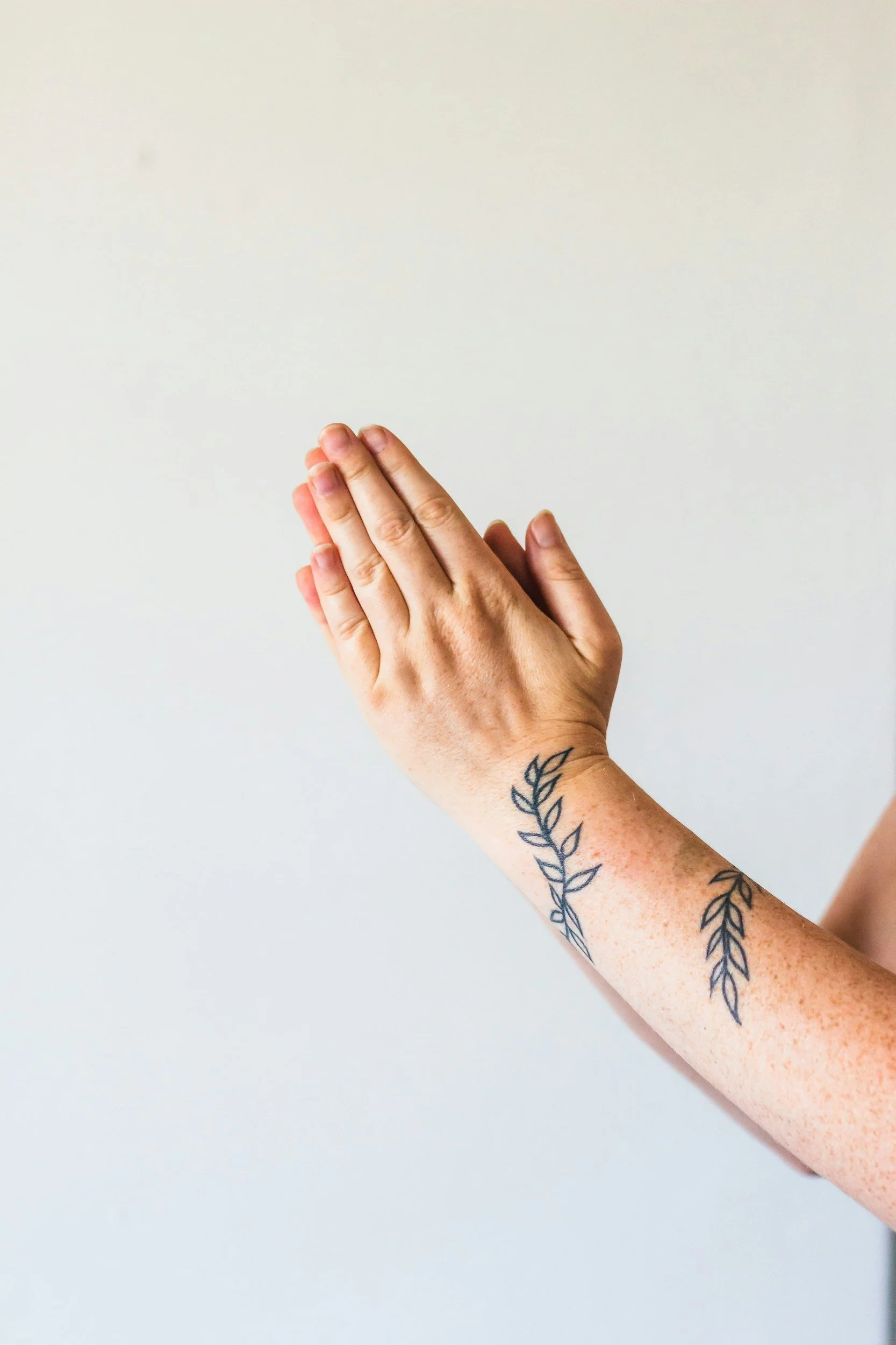 A person's hands are pressed together in a prayer or greeting gesture, with tattoos of leaves on the forearm, against a plain white background.