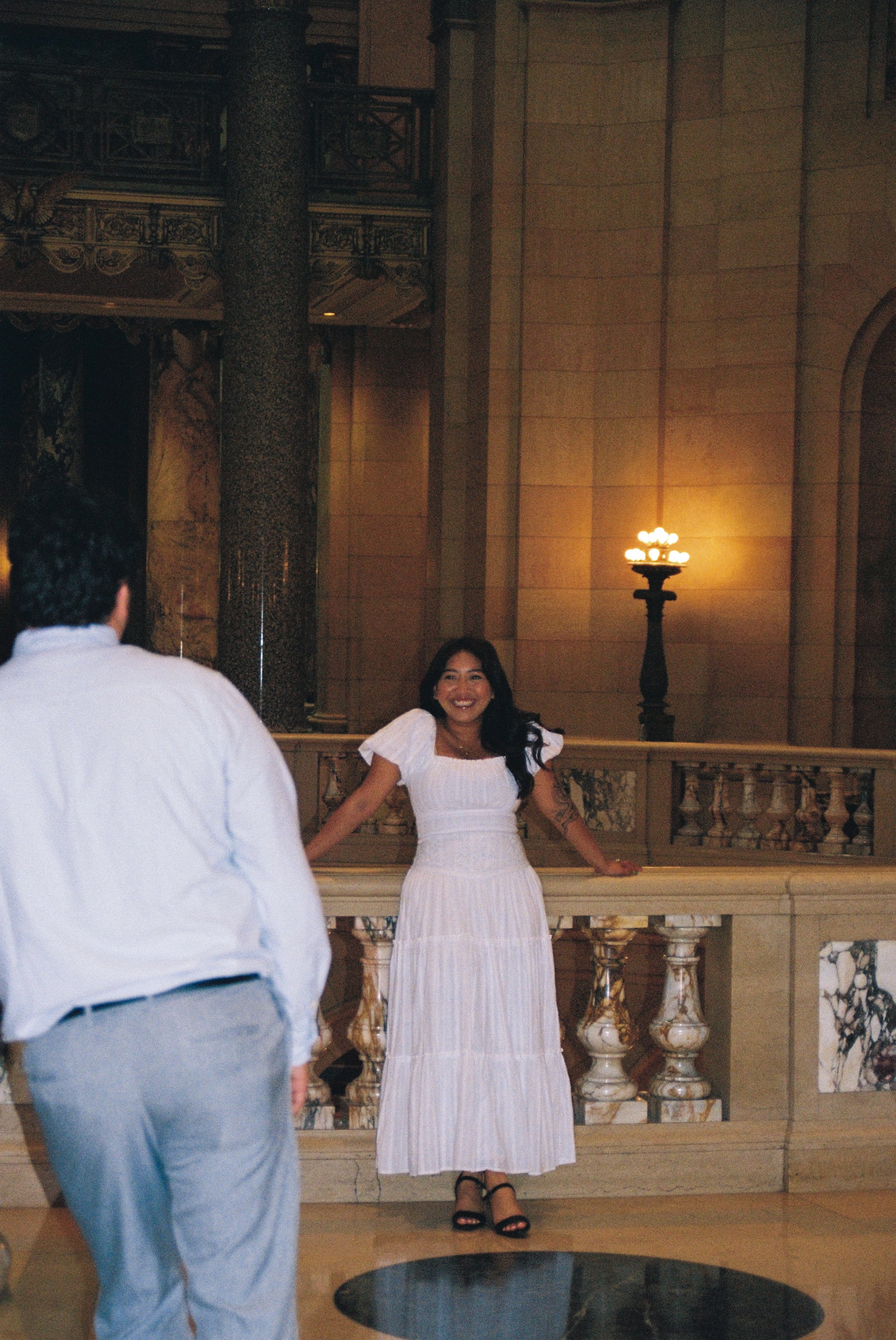 A woman in a white dress standing on a marble staircase inside a grand building, smiling with her hands resting on the railing. A man in a white shirt and gray pants is partially visible in the foreground, facing her. The background features ornate c