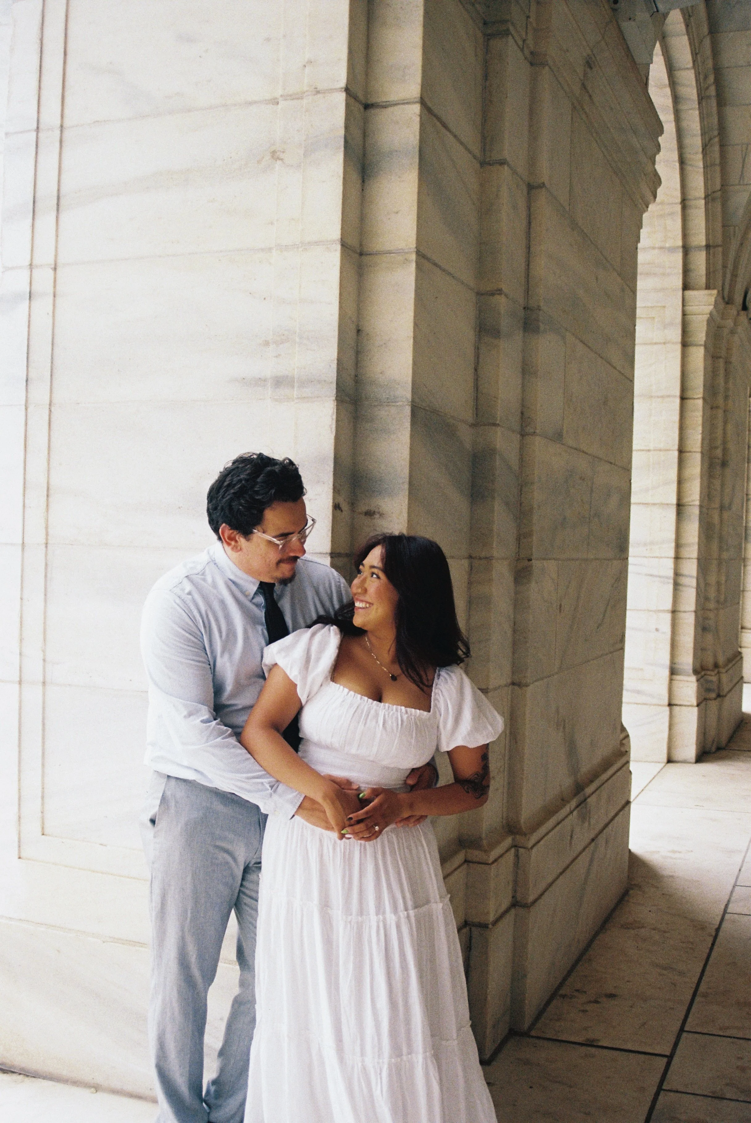 A man and a woman standing close together in front of a stone building, smiling and looking at each other. The man is wearing glasses, a light blue shirt, and grey pants. The woman is wearing a white dress with puffed sleeves and has dark hair.