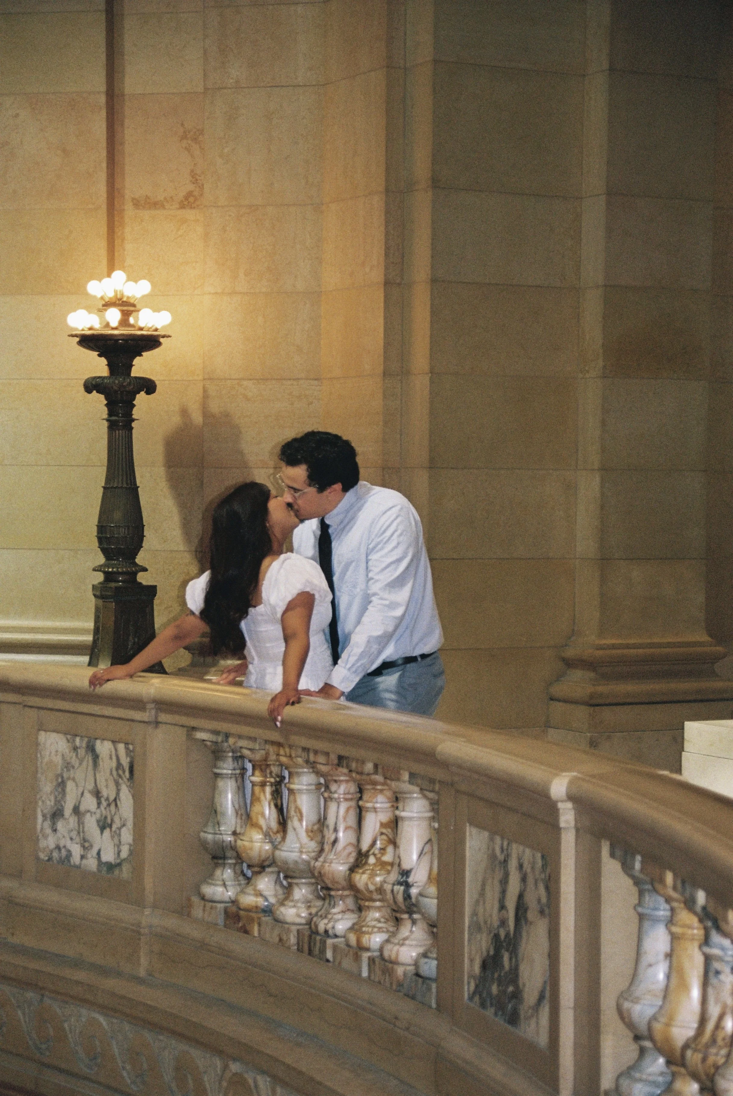 A couple leaning on a marble and wood railing inside a grand building with stone walls and a vintage lamp post.