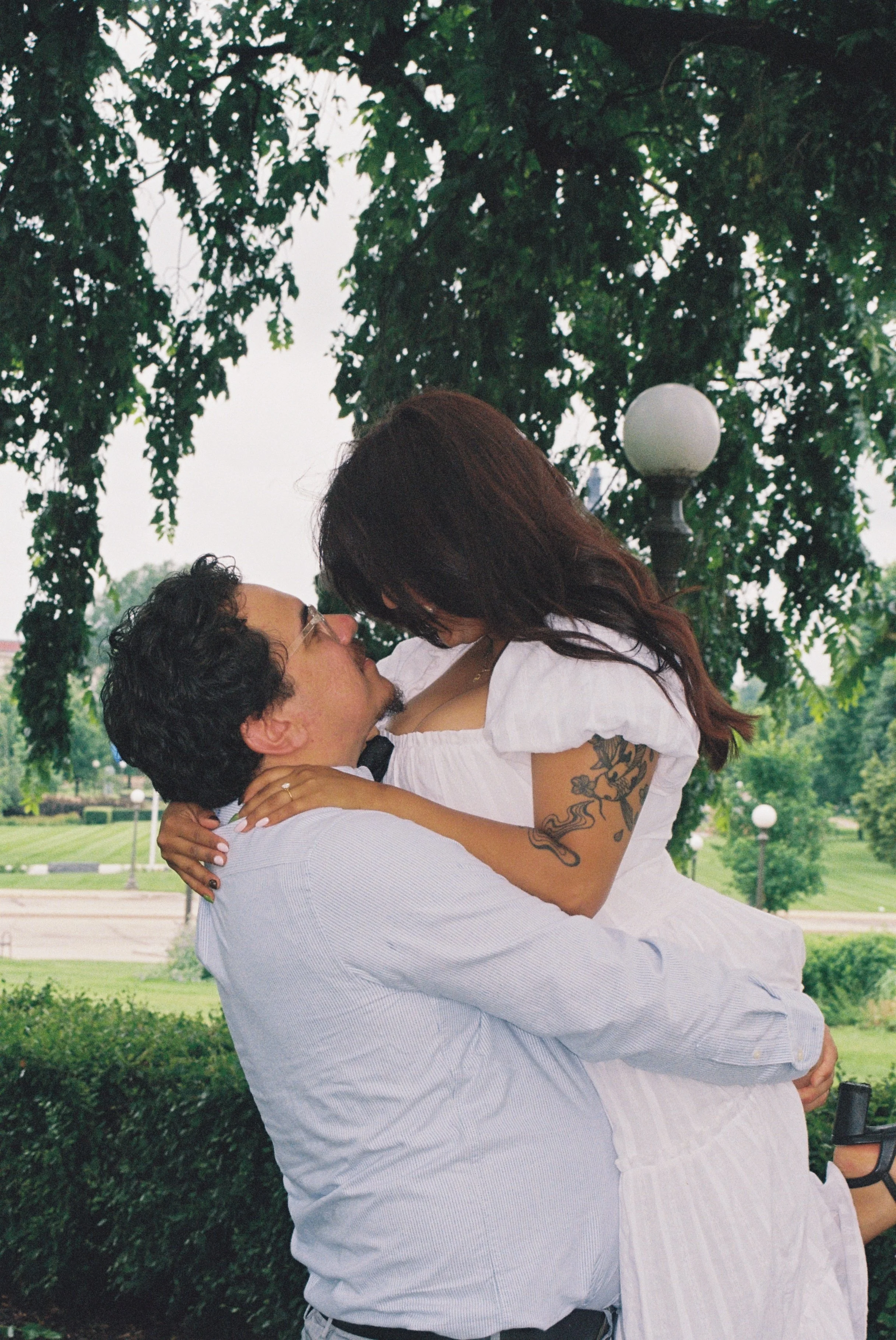 A man and a woman are outdoors, with the man lifting the woman in his arms. They are close together, looking into each other's eyes, under a large tree with green leaves. The woman has long, dark hair and tattoos on her arm, while the man has short, 