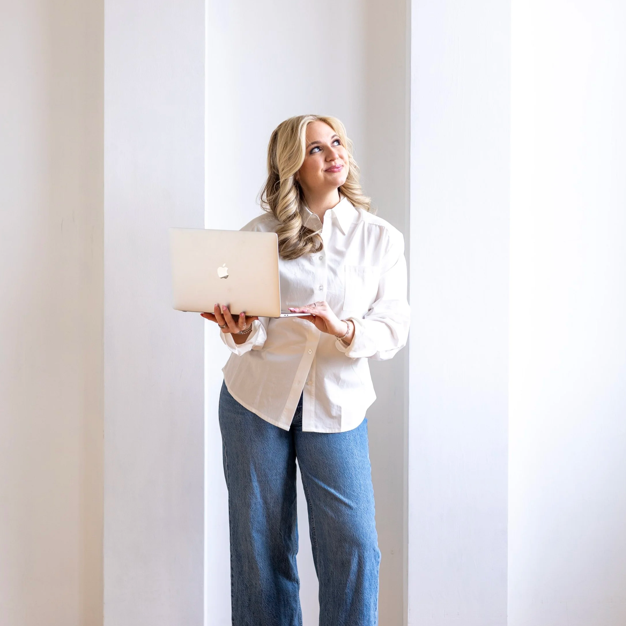 A minimalist workspace with a white desk holding a silver MacBook Air, a pink smartphone, a closed notebook with a floral cover, and a pink folder with glasses on top. Behind, there is a vase of pink roses, four small jars with cork lids, a gold desk lamp, and a framed quote on the wall.