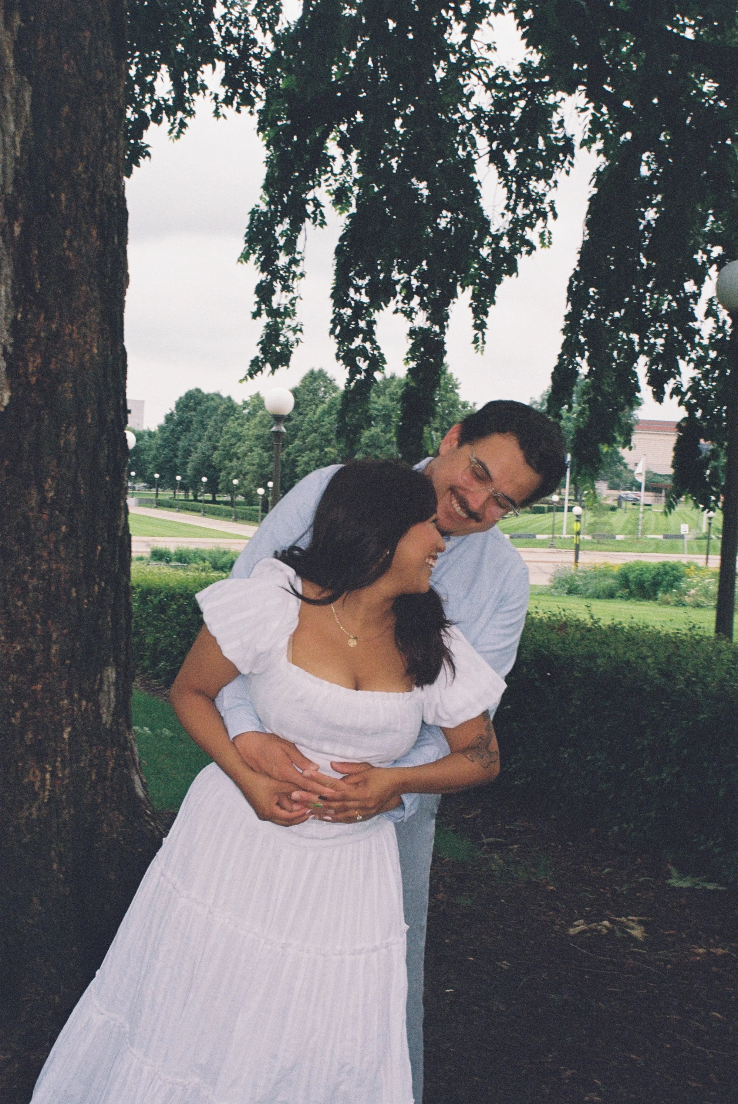 A couple is embracing outdoors in a park with trees and lamp posts, the woman wearing a white dress and the man in a light blue shirt, both smiling and holding each other lovingly.