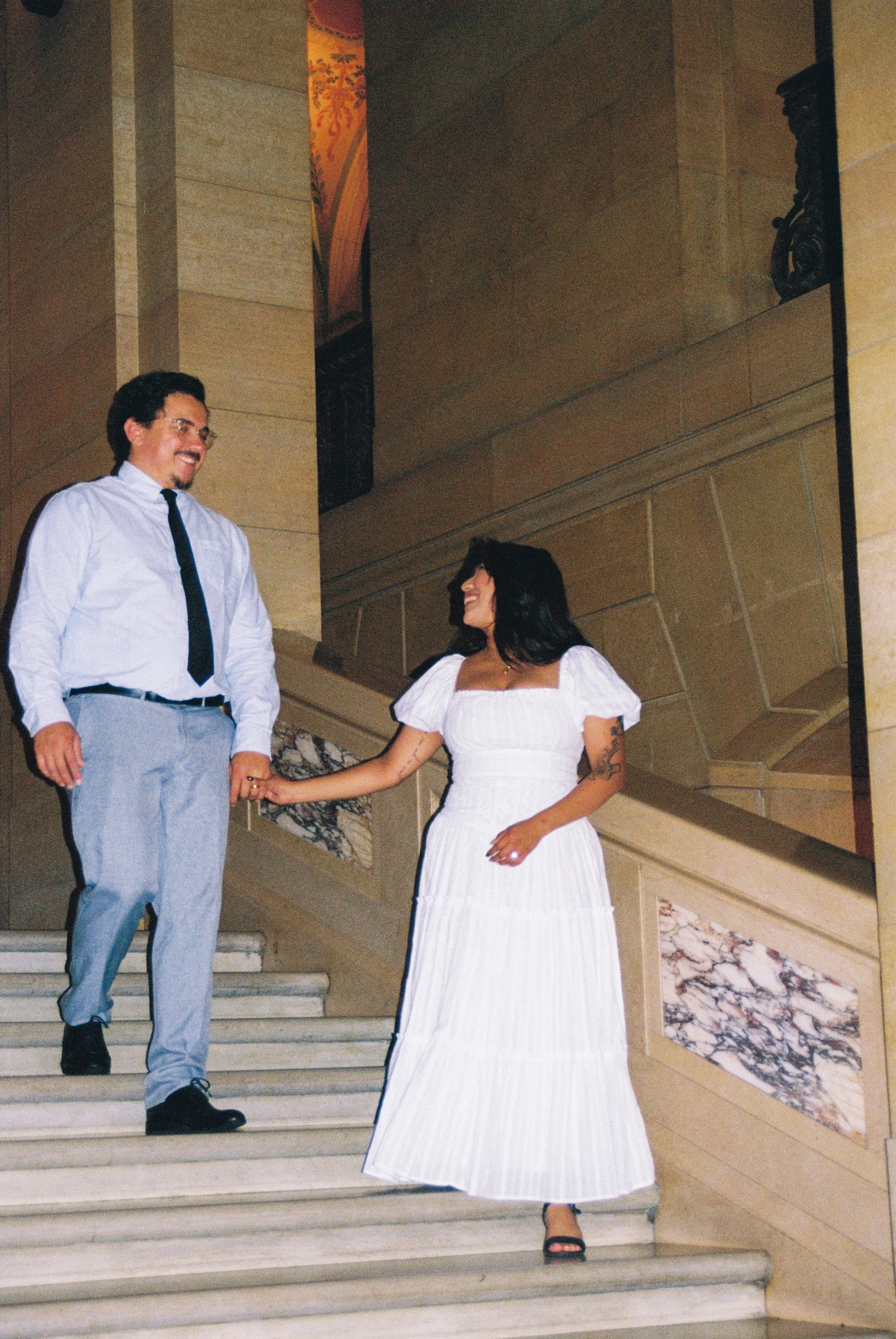 A man and woman holding hands on a staircase inside a building. The man is wearing a white shirt, black tie, and light-colored pants. The woman is in a white dress with black sandals, and has tattoos on her arms. They are smiling at each other.