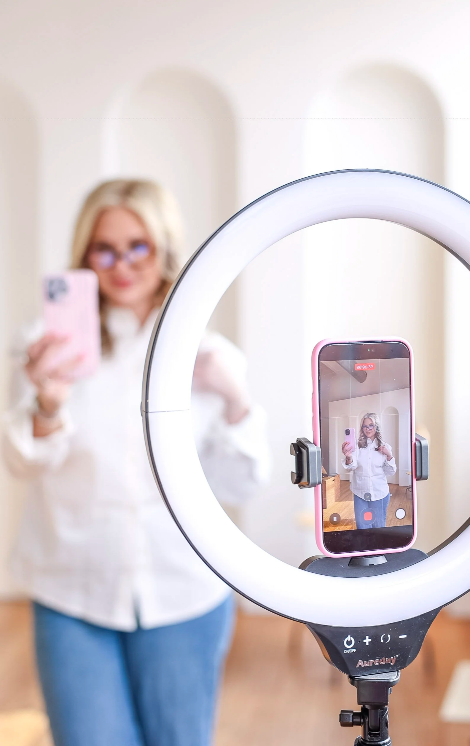 A woman taking a selfie with her phone, visible through a ring light, in front of a mirror.