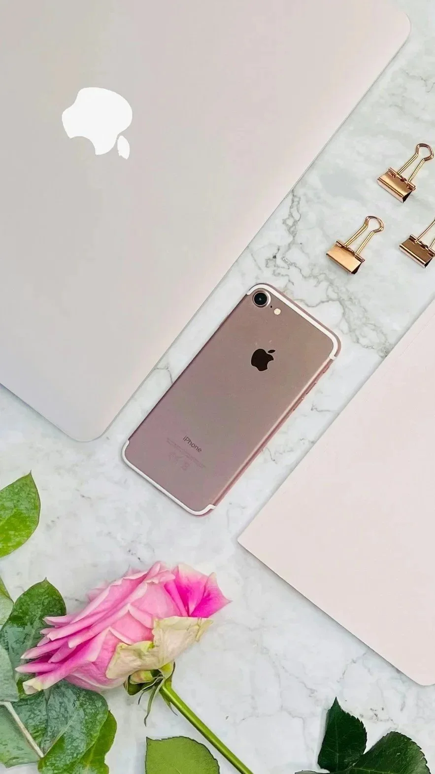 A flat lay of a silver MacBook with an Apple logo, a rose gold iPhone, a pink rose flower, a marble-patterned surface, three gold binder clips, and two pink folders on a white background.