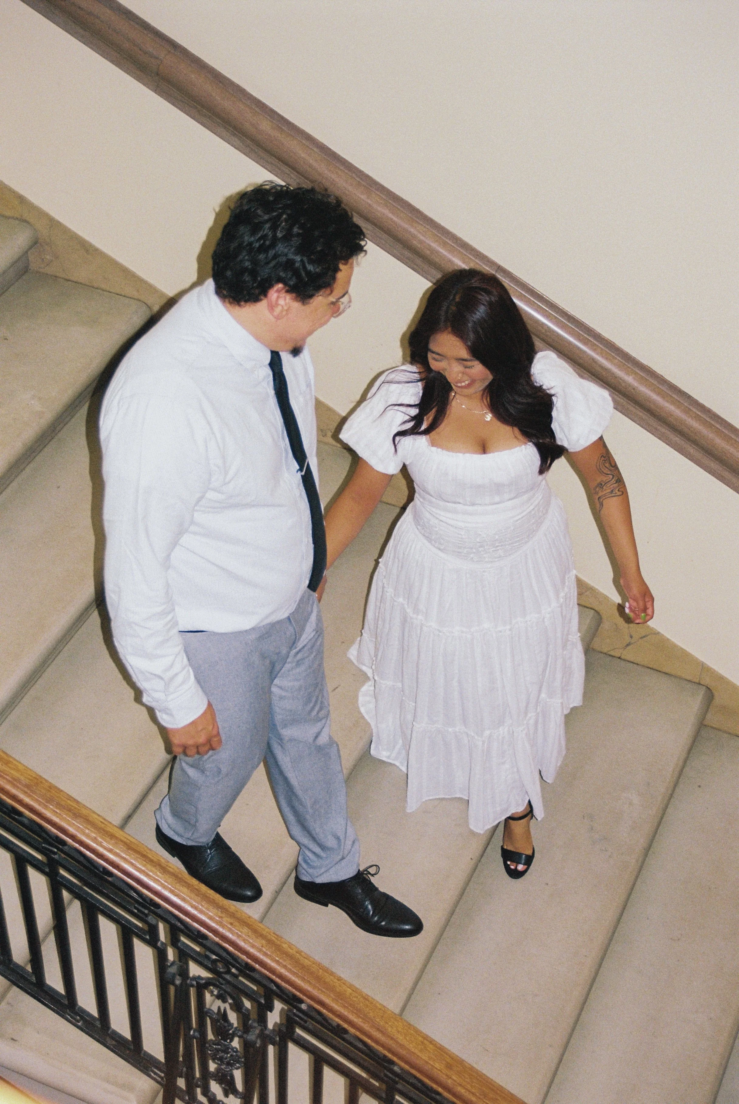 A man and woman are standing on a staircase, smiling and looking at each other. The woman is wearing a white dress, and the man is dressed in a white shirt, gray pants, and a black tie.