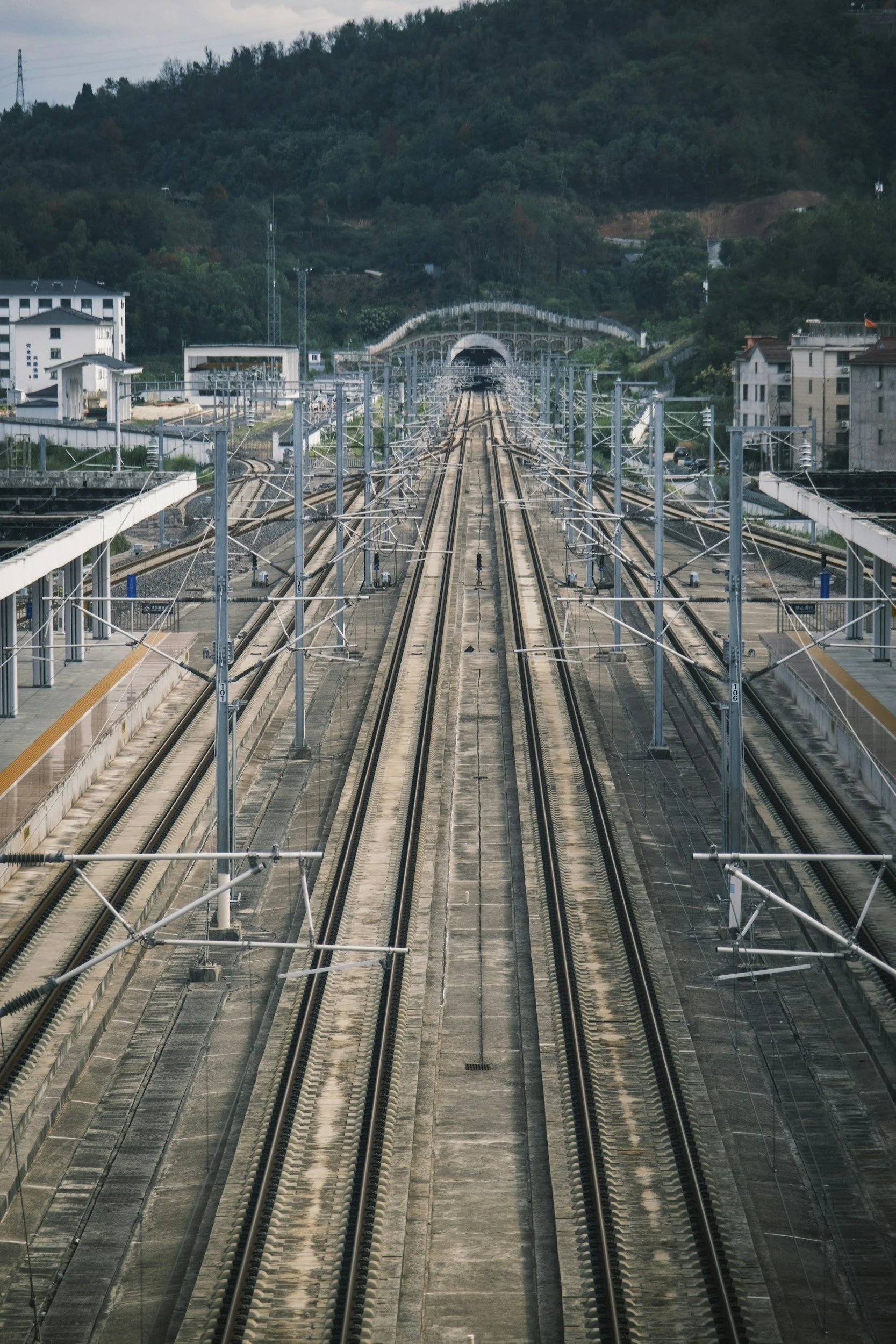 Multiple parallel train tracks extending into a tunnel in the distance, with electrical wires and poles along the tracks, surrounded by buildings and a hilly, wooded landscape.